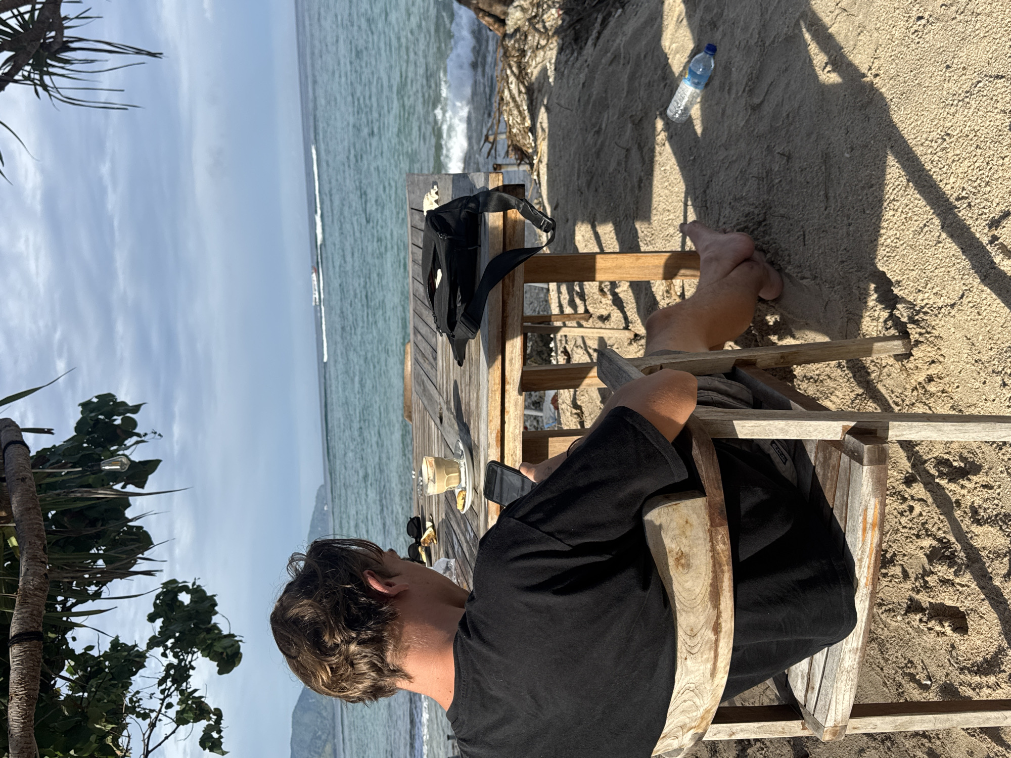 The image depicts an outdoor, sunlit scene at a beachside or coastal location. The primary subject is a person, likely male, with wavy brown hair, wearing a black t-shirt. He is seated in a simple wooden chair, with his back partially to the viewer, looking towards the ocean. His bare feet are visible on the sandy ground beneath the table.
In front of the person is a rustic wooden table. On the table, there is a glass containing a layered drink, possibly an iced latte, accompanied by a small snack on a white plate. A pair of sunglasses and a black smartphone are also on the table. A black sling bag or fanny pack rests on the corner of the table.
The setting features a sandy ground with distinct shadows, indicating a sunny day. A clear plastic water bottle, labeled "Aqua", lies on the sand near the table. To the left of the frame, tropical-looking foliage, possibly from a palm tree or similar plant, provides some natural framing. In the background, a vast expanse of blue-green ocean with breaking waves stretches towards the horizon under a light blue sky with scattered clouds. In the distance, faint outlines of land or islands can be seen.
The activity suggests a moment of relaxation, possibly enjoying a drink and snack while taking in the ocean view. The strong shadows imply it's daytime, likely mid-morning or late afternoon. The overall atmosphere is tranquil and leisurely. A small tag with the letters "AOQ" is faintly visible on the person's shorts or bag.