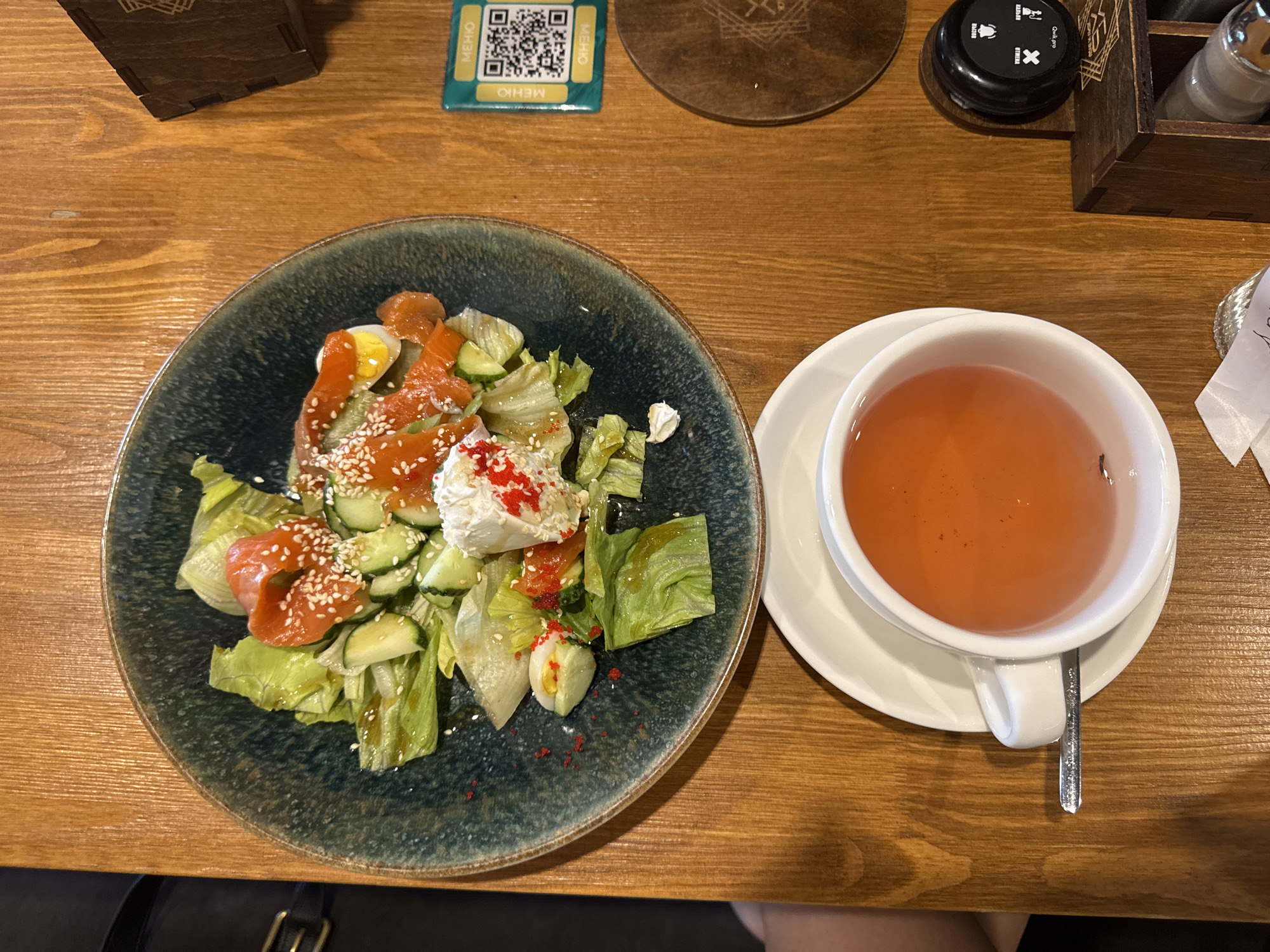 This is a close-up, top-down shot of a meal on a wooden table, likely in Kazan, Russia, given the presence of a QR code for "MEHIO" which translates to "menu" in Russian. The scene features a dark, textured plate filled with a salad. The salad includes salmon, cucumber slices, lettuce, and a dollop of white cheese topped with red roe and sesame seeds. To the right of the salad is a white teacup filled with a reddish-orange liquid, placed on a matching saucer. A silver spoon rests on the saucer. In the background, a wooden box contains salt and pepper shakers. Also visible is a black call button and a circular wooden coaster. The lighting suggests an indoor setting. There are no people or visible actions in the image.