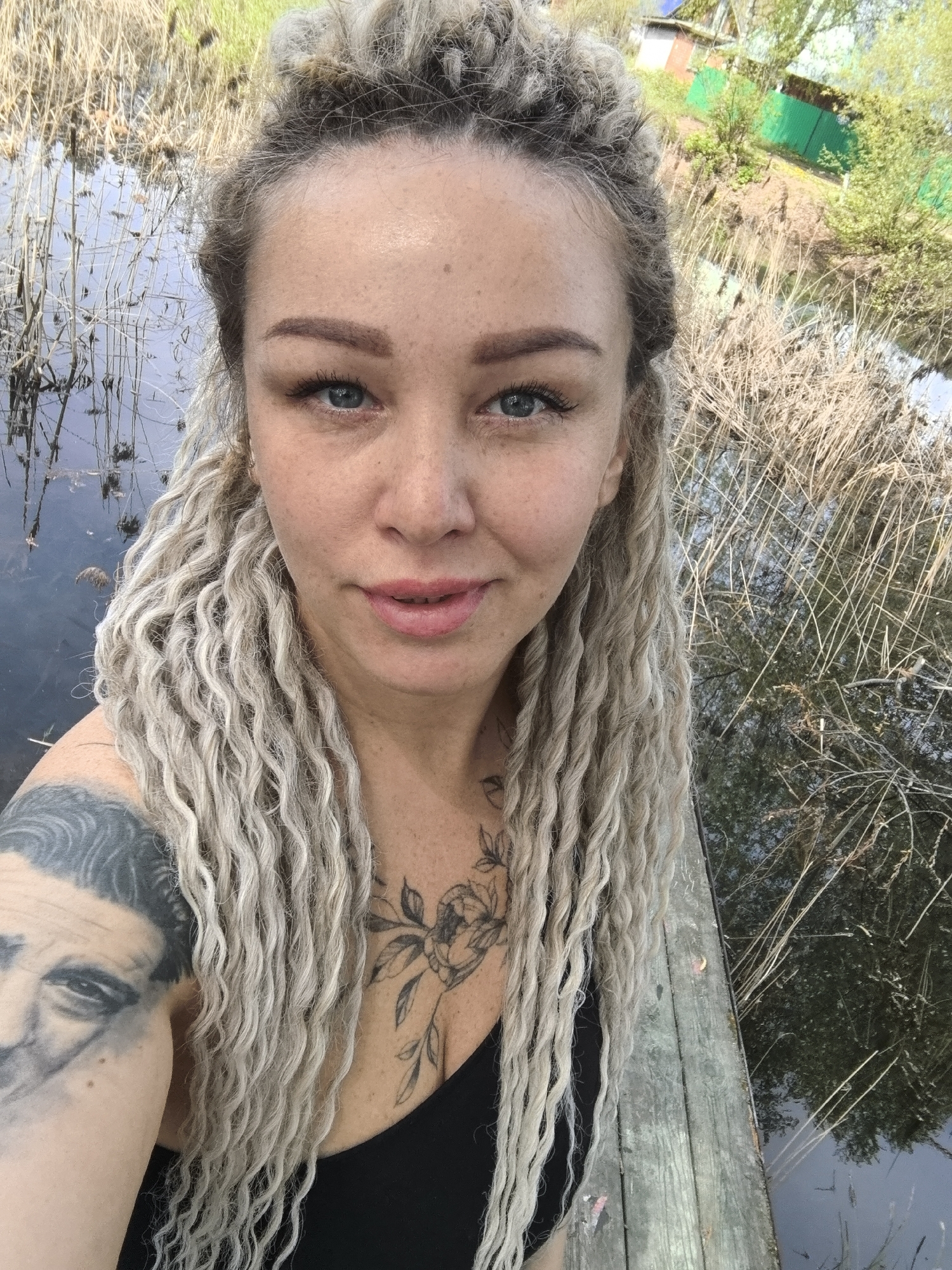 A woman with dreadlocks and tattoos poses for a selfie outdoors. She has light skin, blue eyes, and light pink lips. Her hair is blonde and styled into dreadlocks, with some gathered on top of her head. Visible on her left shoulder is a tattoo of a face. On her chest, there is a floral tattoo. She wears a black top.
The background shows a marshy area with tall reeds and water. In the distance, there are green trees and what appears to be a building with a green fence. The ground she stands on looks like a wooden deck or platform. The location is presumed to be near Noviy, Russia based on the request context.