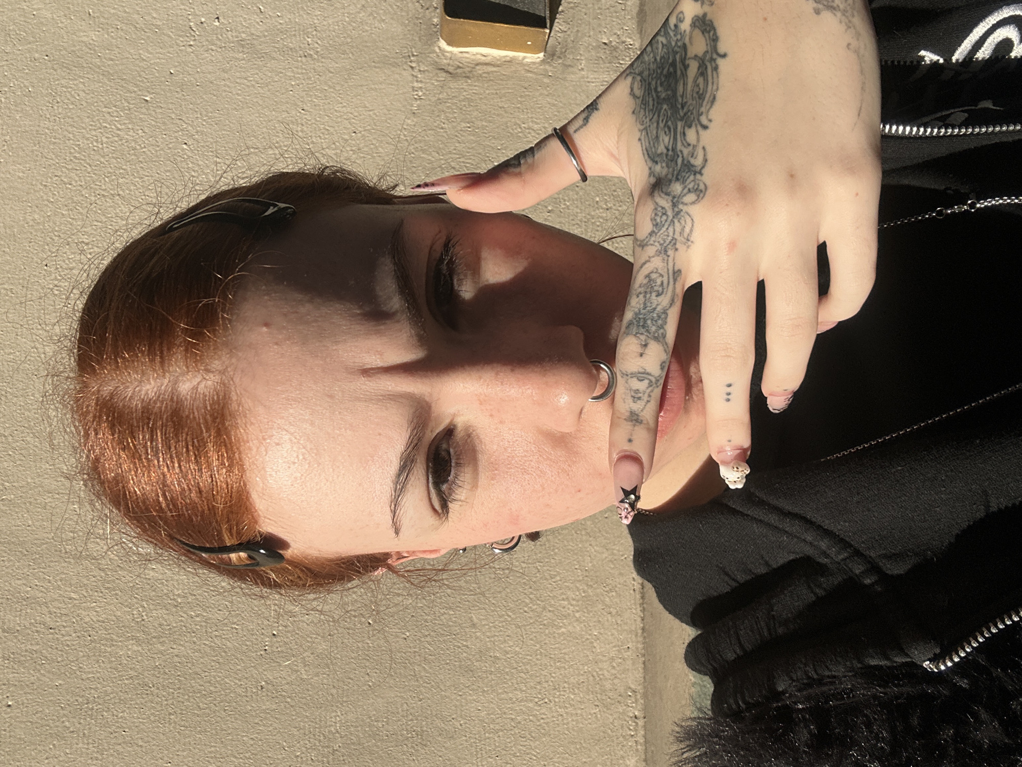 The image is a rotated, close-up portrait of a person with auburn hair and fair skin, illuminated by bright sunlight. The subject's right hand, heavily tattooed with intricate black line art patterns across the back and fingers, is positioned near their face. A simple silver band ring is worn on the thumb of the tattooed hand. The fingernails are long and manicured; the index finger displays a black star design, and the pinky finger features a small, white, three-dimensional charm, possibly a skull.
The person wears black hair clips, a septum piercing, and a nostril ring. Their eyes are slightly squinted due to the direct light, and visible freckles dot their skin. They are dressed in a black zippered hoodie or jacket, with a silver chain necklace visible beneath. The garment appears to have a textured, possibly furry, trim.
The background consists of a plain, light-colored, textured wall, indicating an outdoor setting. Strong, direct sunlight creates prominent shadows on the person's face and clothing, suggesting it is daytime. A small, metallic, rectangular object is partially visible in the upper background. There is no visible text in the image.