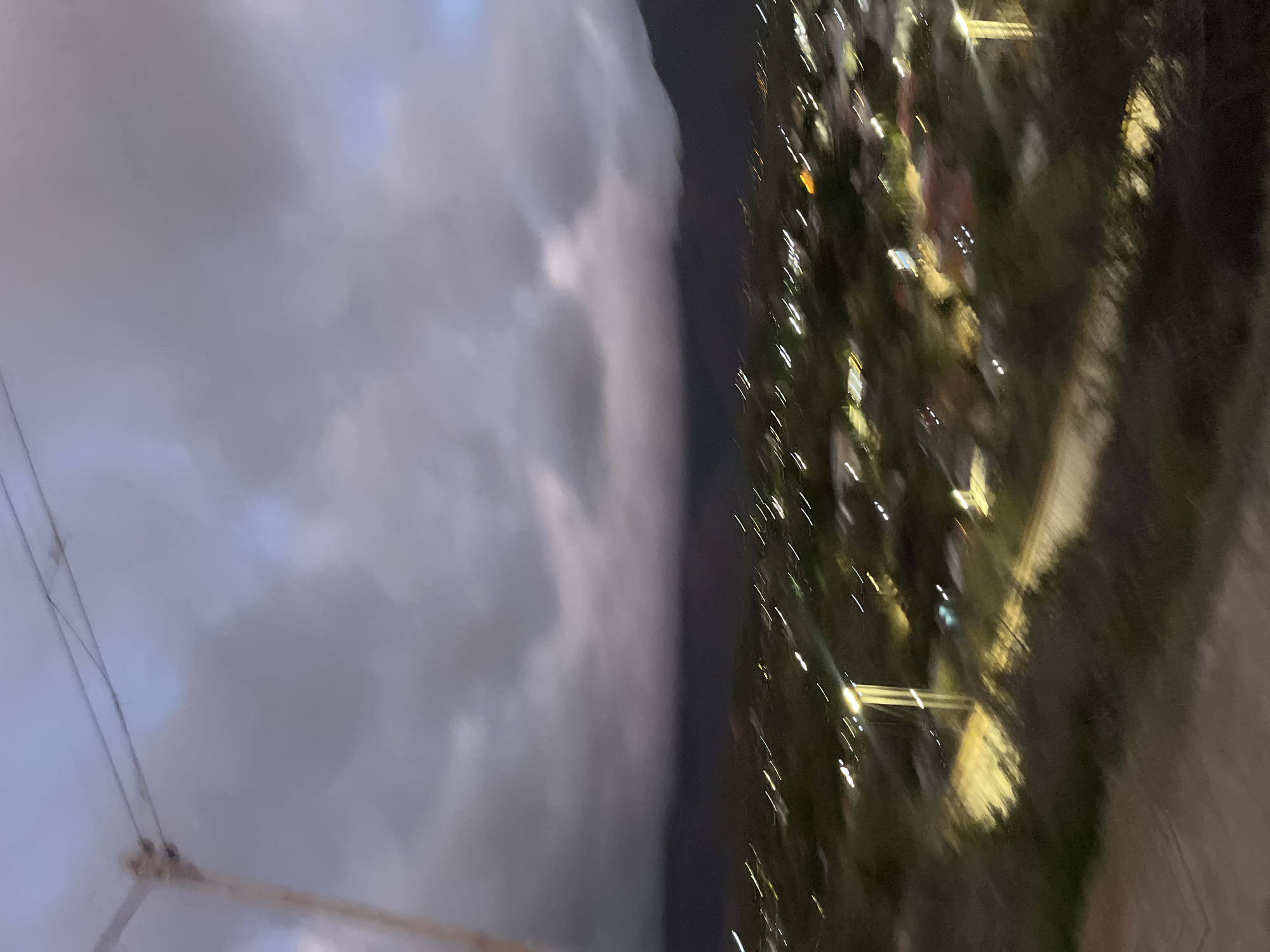 The image shows a blurred aerial view of a city at dusk. The sky is filled with dark, heavy clouds, with hints of blue and pink light breaking through. Below the clouds, dark mountains form a silhouette against the sky. The city lights twinkle in streaks across the landscape, indicating movement or a long exposure. Buildings and vegetation are visible, but are indistinct due to motion blur. A metal pole and wires are present in the upper left corner of the image. The scene is likely Santa Ana, Venezuela, given the context provided.