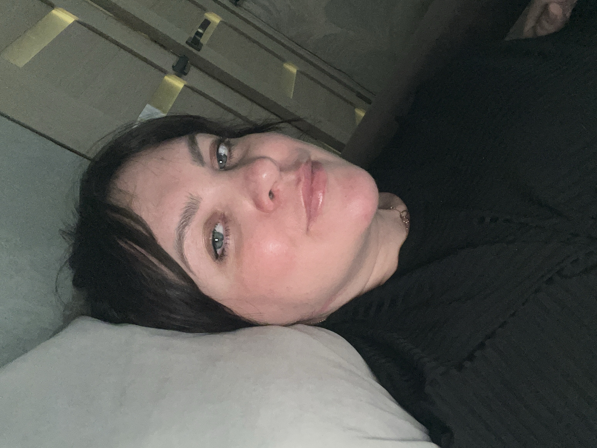 A woman with dark hair and fair skin is lying on a light-colored pillow. She is wearing a dark, possibly black, garment. The background appears to be a wall or headboard with decorative elements, possibly illuminated. The image is taken at an angle, as if the camera is held sideways. There is no indication of the location being Noviy, Russia.
