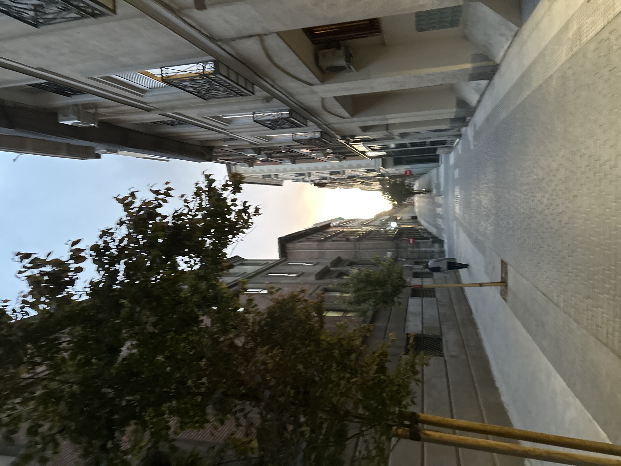 The image depicts a street view in Madrid, Spain. The perspective is looking down a narrow street lined with buildings on both sides. The buildings are predominantly light-colored with architectural details such as window boxes and balconies. A tree with green leaves is visible on the left side of the frame. The street appears paved with light-colored stones or tiles. There is a person walking on the right side of the street. The sky is visible in the distance at the end of the street, suggesting either sunrise or sunset. A scaffolding is along the right sidewalk.