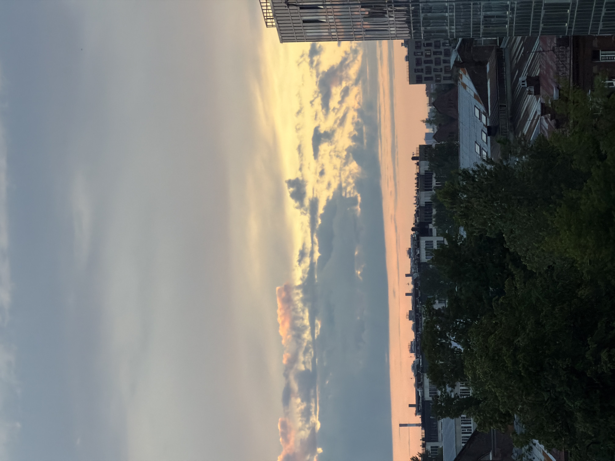 This is a high-angle shot of a cityscape at dusk in an unknown location in Russia. The sky is filled with dramatic clouds, with streaks of pink and orange light from the setting sun breaking through. In the foreground, the dark silhouettes of trees are visible, obscuring some of the buildings below. These buildings appear to be a mix of older structures with pitched, rusty roofs and newer, modern glass-fronted buildings. The overall impression is one of an urban environment transitioning into nighttime. There are no people or specific actions depicted in the scene.