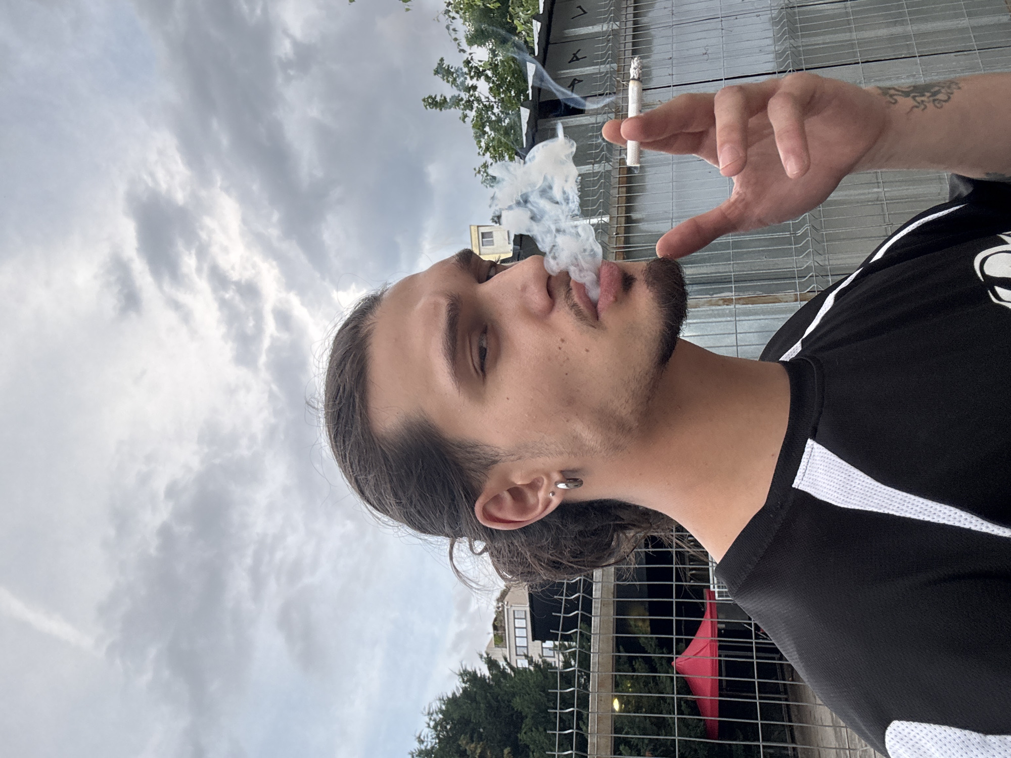 A young man with long, dark hair tied back and a goatee is shown in profile, smoking a cigarette. He is wearing a black t-shirt with white accents. He has a hoop earring in his left ear and a tattoo on his right forearm. The background features a cloudy sky, trees, buildings, and a grey metal fence. The location is identified as being in the city of Sofia, Bulgaria.