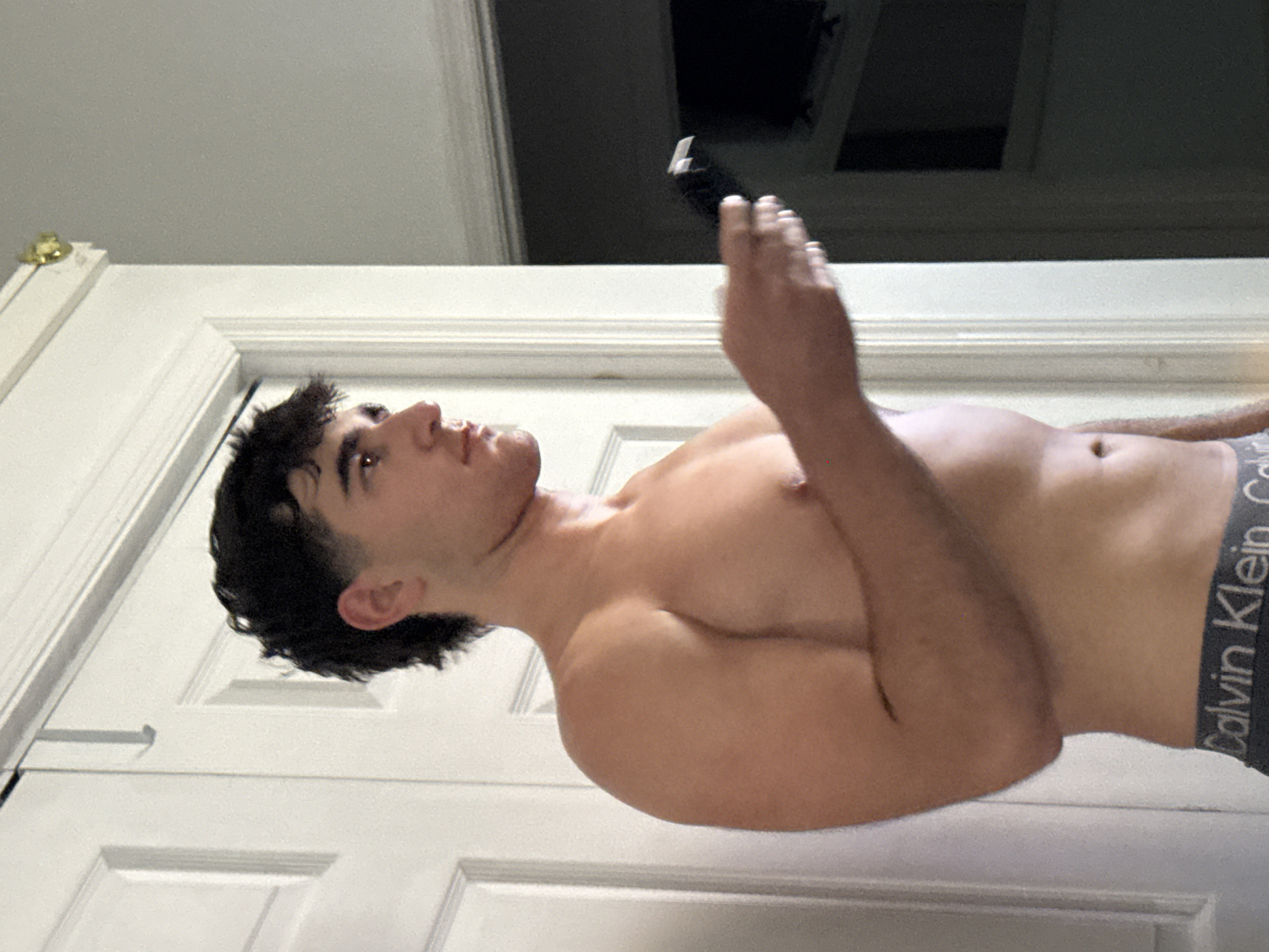 A young man, shirtless and wearing Calvin Klein underwear, is standing indoors, holding a black electric trimmer in his right hand. He is looking to his right, with his hair styled in a modern mullet. The background consists of a white doorframe with paneling, and a dark doorway leading to another room where a television is visible. The image appears to be a personal photograph taken in a domestic setting in Bellmore, United States of America.