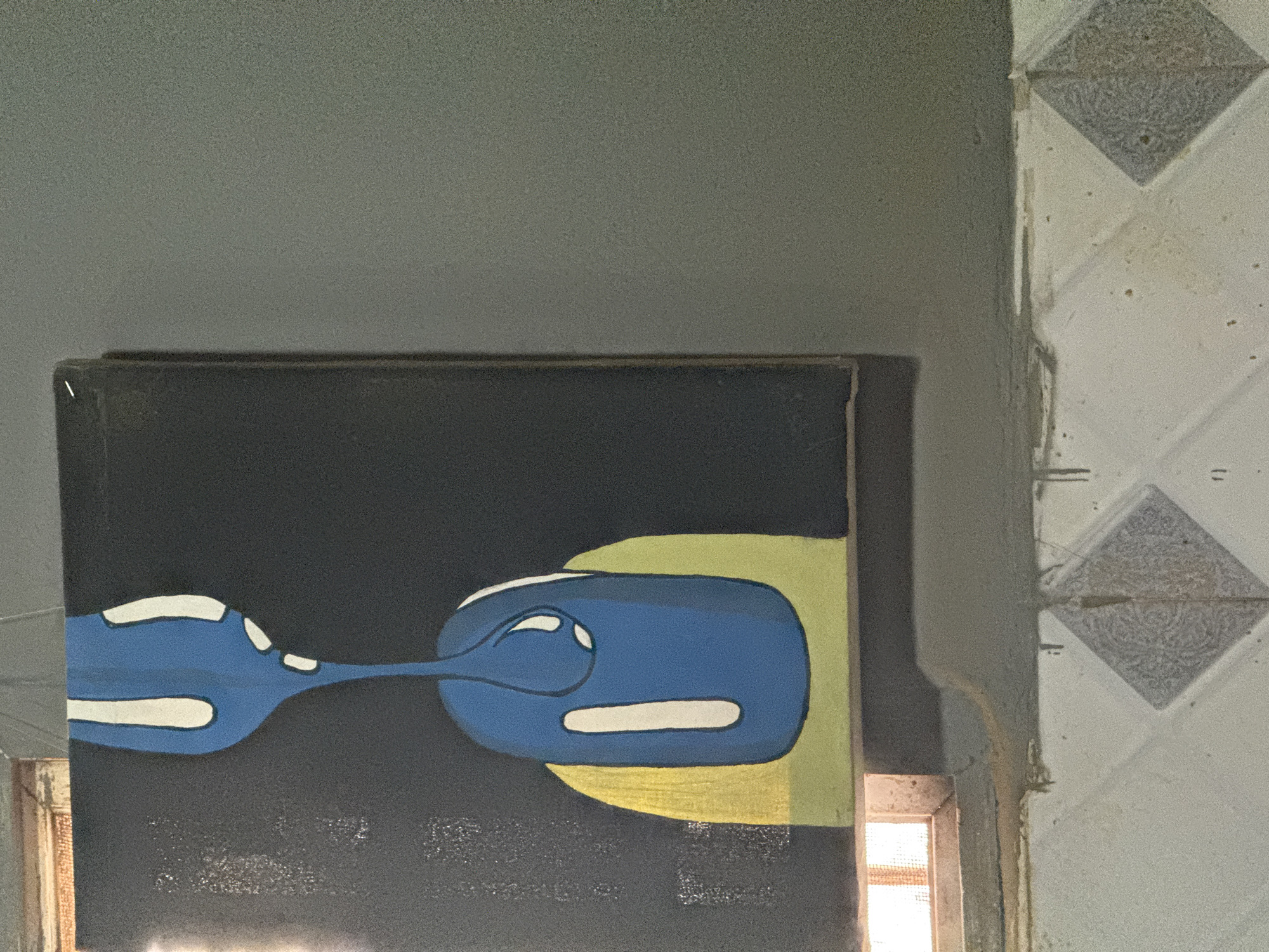 The media file captures an indoor scene dominated by a horizontally oriented painting against a dark background. The artwork features abstract, stylized shapes primarily in blue with white outlines and highlights, resembling interconnected forms that evoke the appearance of computer mice or elongated organisms. To the right of these blue elements, a rounded, yellowish-green area completes the painted composition.
Above the painting, a plain, light grey wall with some texture is visible. To the right of the painting, a different wall section features light-colored, possibly cream or off-white, patterned tiles. These tiles display a square or diamond design with a textured, almost swirled pattern within them. The corner where the walls meet appears somewhat unfinished or aged.
A strong, bright light source emanates from the bottom right of the image, below and to the right of the painting, suggesting an open window or doorway to the outside. This light casts a glow on the adjacent surfaces. The scene is static, depicting the art piece in what appears to be a rustic or simply adorned interior setting. There are no people visible, no discernible activity taking place, and no visible text in the image. The bright illumination indicates it is daytime. The overall image has a noticeable grainy quality.
