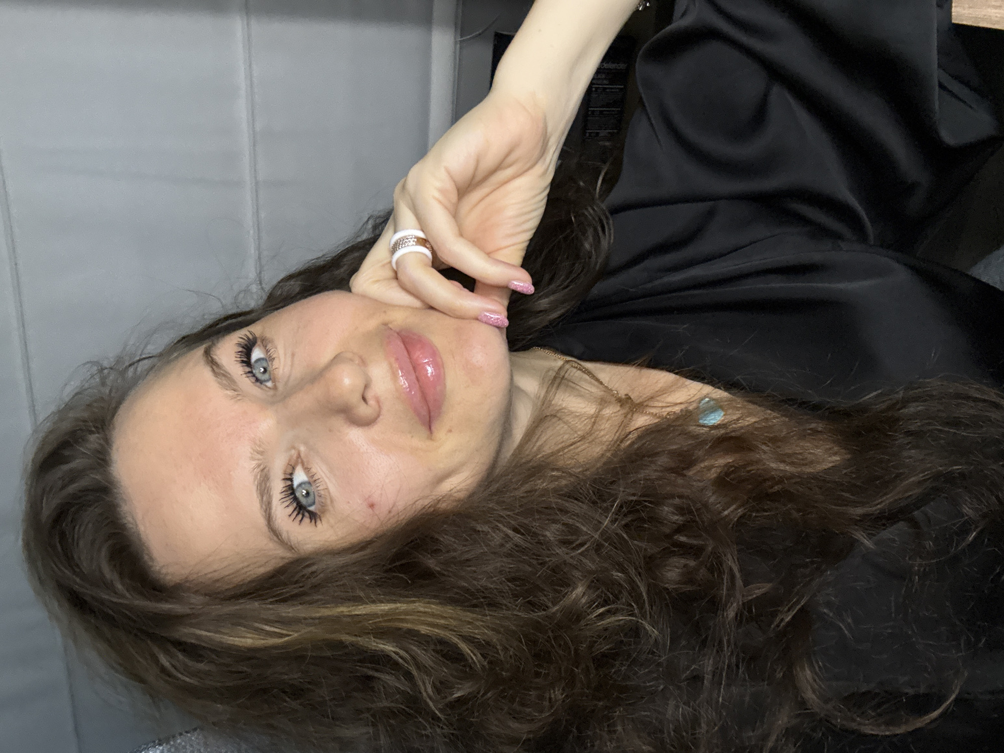 A woman with shoulder-length brown hair and blue eyes is posing for a selfie. She is wearing a black silk robe or shirt. She has on a pink lip gloss and mascara. She has rings on her finger with pink, white, and gold details. Her hand is touching her face. The location is not discernible from the image.
