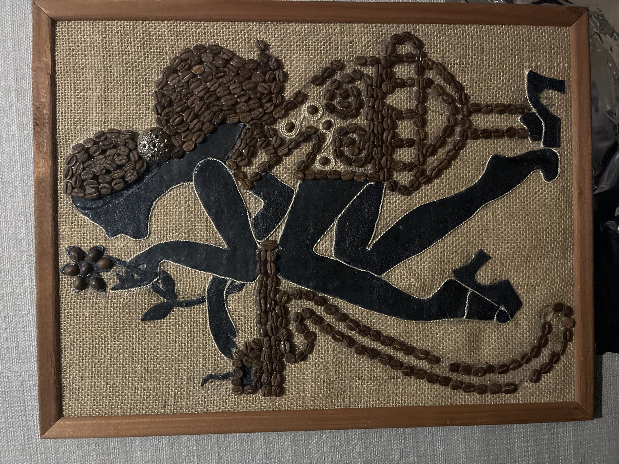 The image shows a framed artwork created from coffee beans. The artwork is a silhouette of a woman in a dress, holding a flower. The woman's hair and parts of her dress are made from coffee beans, while her silhouette is cut from black material. The artwork is displayed on a burlap-like background and is framed in wood. The location of this artwork in Saratov, Russia is not discernible from the image.