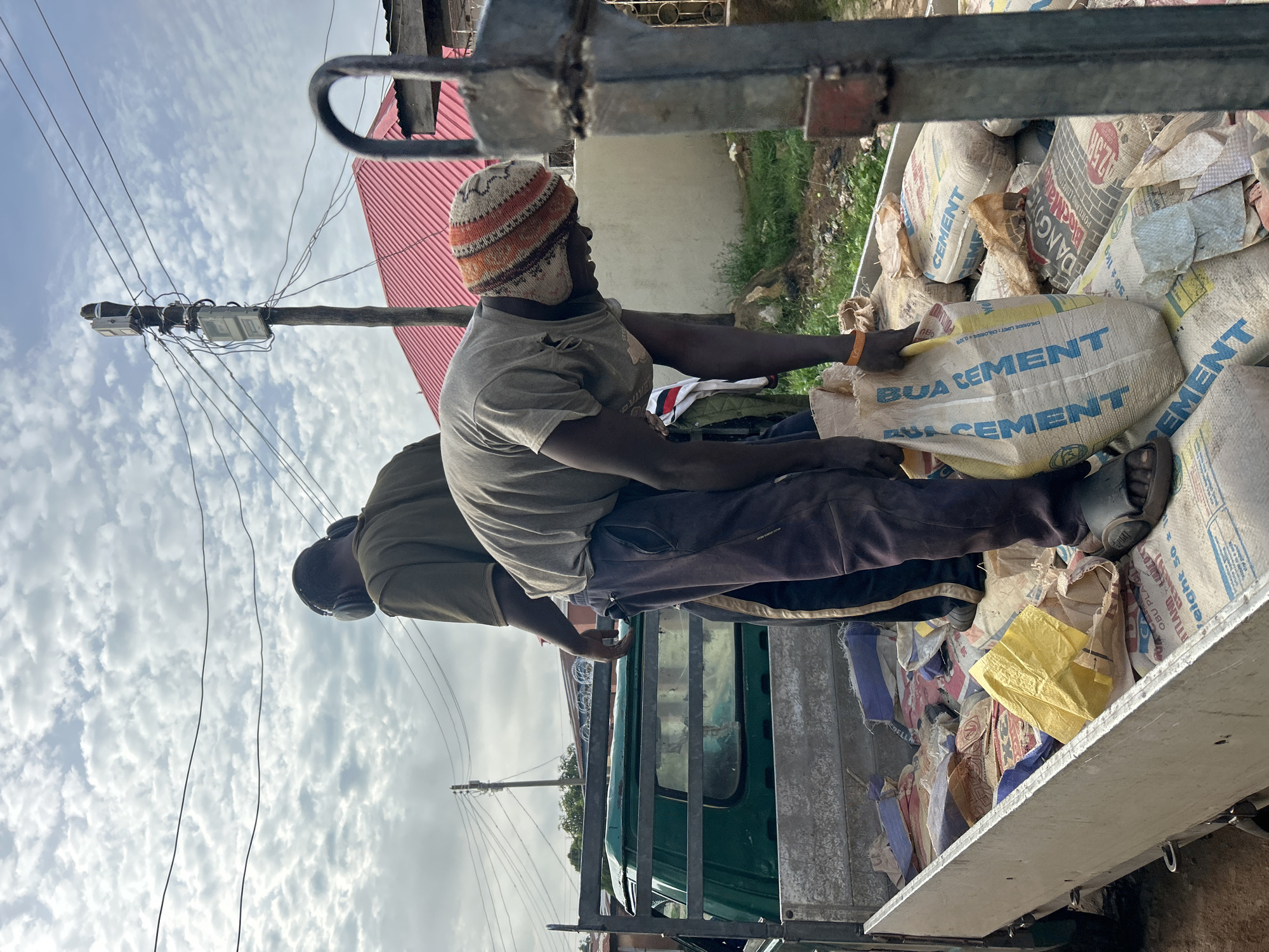 In Jos, Nigeria, two men are in the bed of a green truck filled with bags of cement. One man, wearing headphones and a green shirt, stands to the left. The other man, wearing a knitted cap, is bent over and holding a yellow and blue bag labeled "BUA CEMENT." The truck is parked outdoors with a cloudy sky overhead. There are power lines and a utility pole visible in the background, as well as a red-roofed building. The scene appears to be a construction or delivery site.