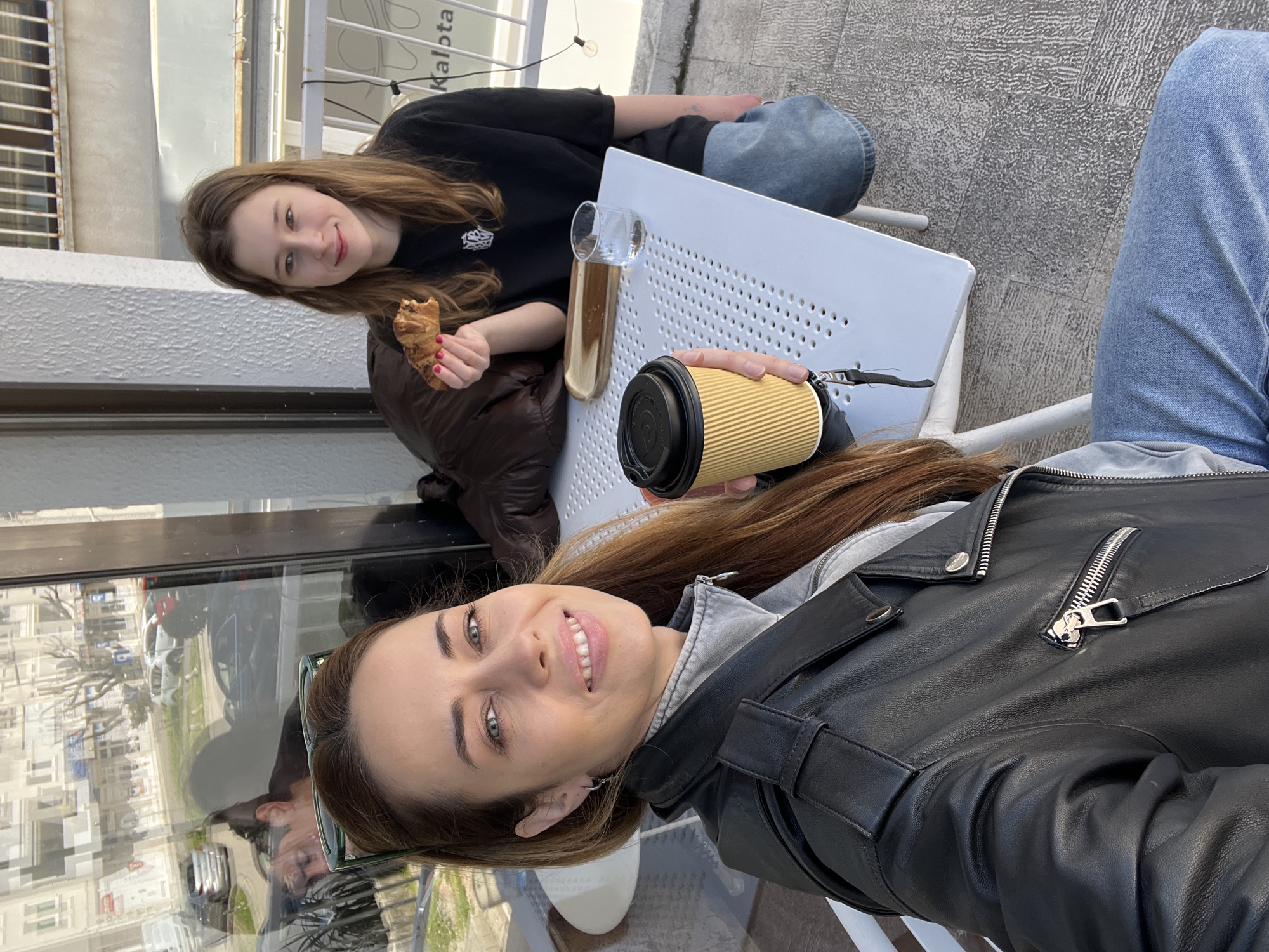 Two women are at an outdoor cafe in Bar, Montenegro, sitting at a white metal table. One woman, wearing a black leather jacket over a grey hoodie and light-wash jeans, holds a coffee cup. The other woman, dressed in a black shirt and jeans, holds a croissant. A tall glass with a liquid inside is placed on the table. The cafe is near a street with cars parked along it. A building is reflected in the window behind them.