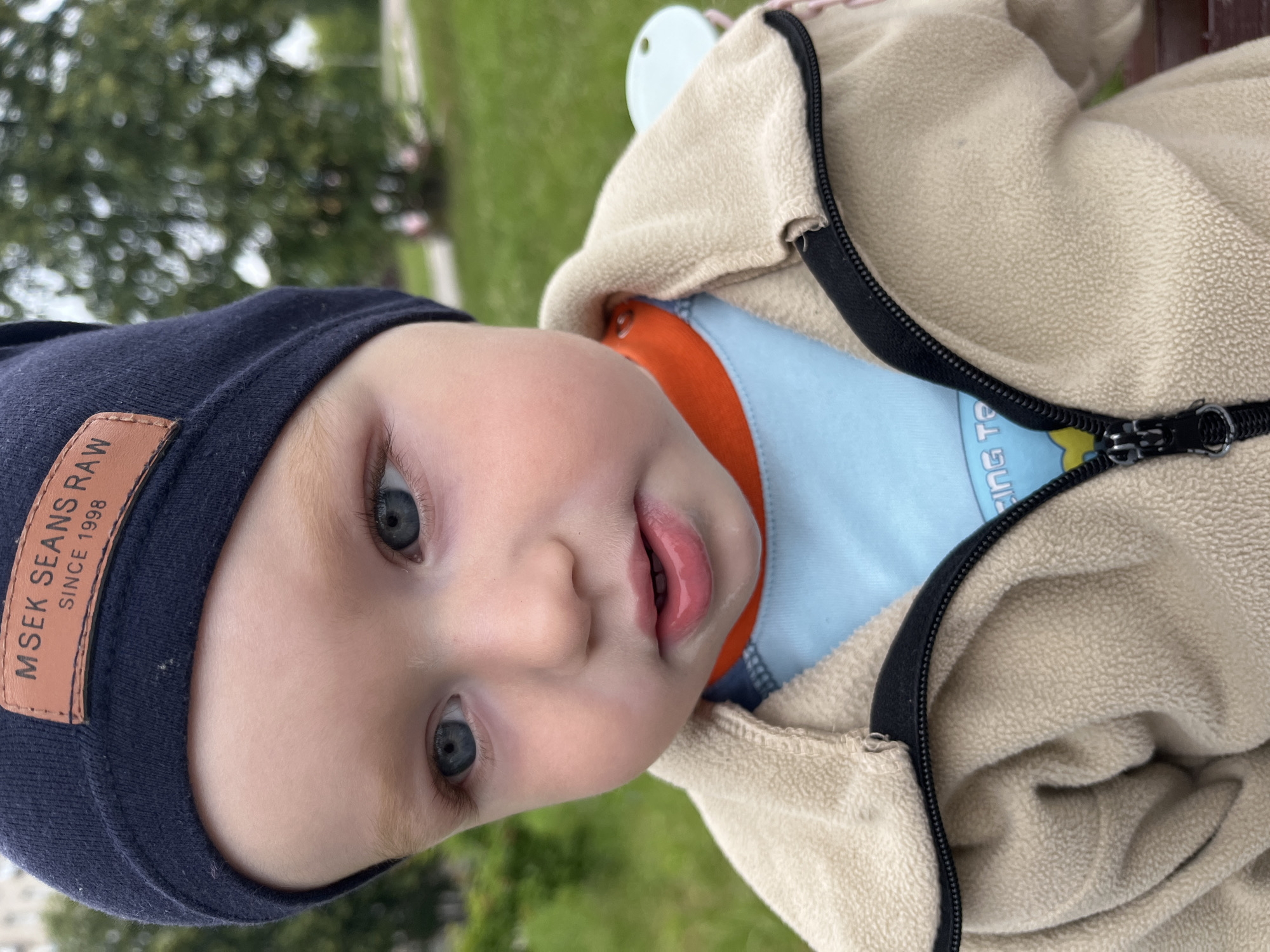 A close-up, eye-level, outdoor medium shot features a young child wearing a navy blue beanie with a brown leather tag that reads "MSEK SEANS RAW SINCE 1998." The child has light-colored hair and blue eyes, and is looking slightly off-camera to the right. The child is wearing a light blue shirt with an orange collar, layered under a beige zippered fleece jacket. The background is blurred, showing green grass and trees, suggesting a park or outdoor recreational area in Chaikovsky, Russia. The lighting is natural daylight.