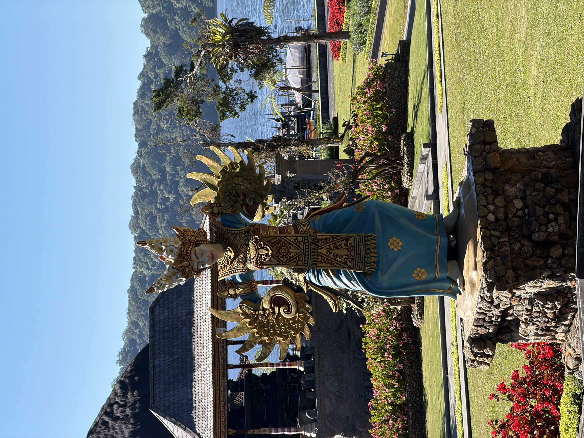 A statue of a Balinese dancer stands on a stone pedestal in a park. The statue is adorned in traditional attire, with a golden headdress and elaborate golden ornaments on its chest and arms. The dancer's costume is primarily blue with golden accents. In the background, a traditional Balinese building with a thatched roof is visible, along with a body of water, likely a lake, bordered by lush green hills covered in trees. The park features well-maintained green lawns and vibrant flower beds in shades of pink and red. The sky is clear and blue, indicating a sunny day. The location is identified as Baturiti, Indonesia.