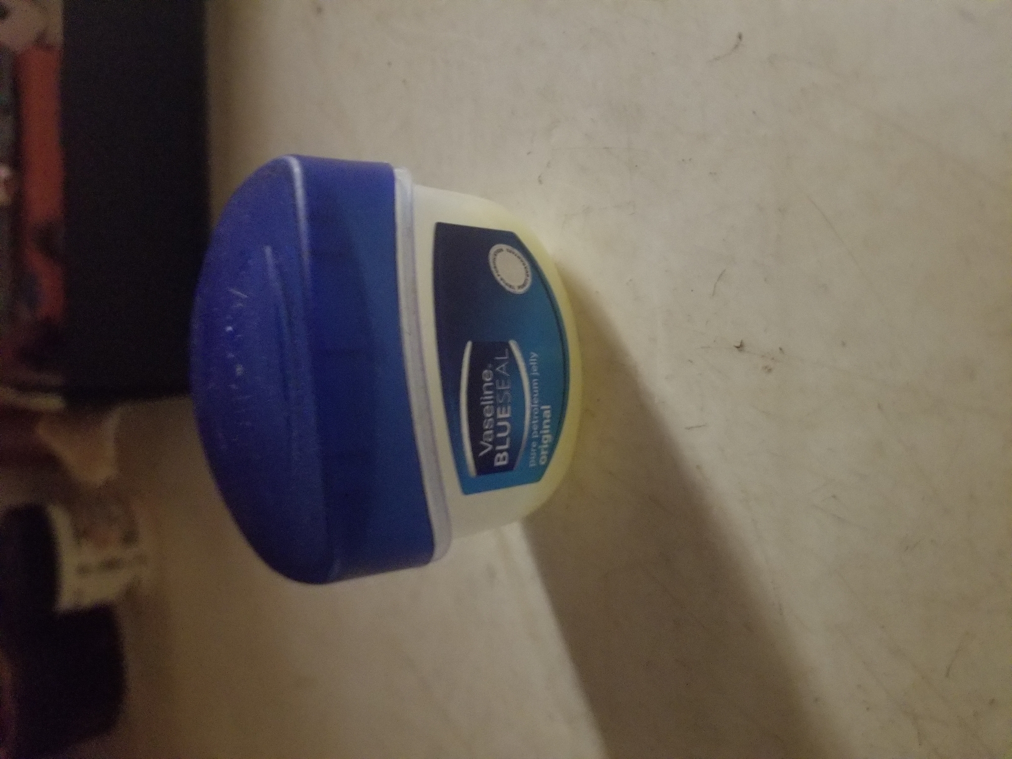 A small, white, plastic jar of Vaseline Blue Seal petroleum jelly, with a blue lid, sits on a light-colored, textured surface. The label on the jar is partially visible and reads "Vaseline BLUE SEAL" in blue text, with "Pure petroleum jelly original" below it in smaller text. A faint shadow falls across the surface to the left of the jar, suggesting a light source from the right. The background is blurred and indistinct, showing dark shapes and lighter, out-of-focus objects. There are no people or discernible actions taking place in the image. The overall scene is domestic and still. There is no information that allows for the determination of a specific location within Jauro Yinu, Nigeria.