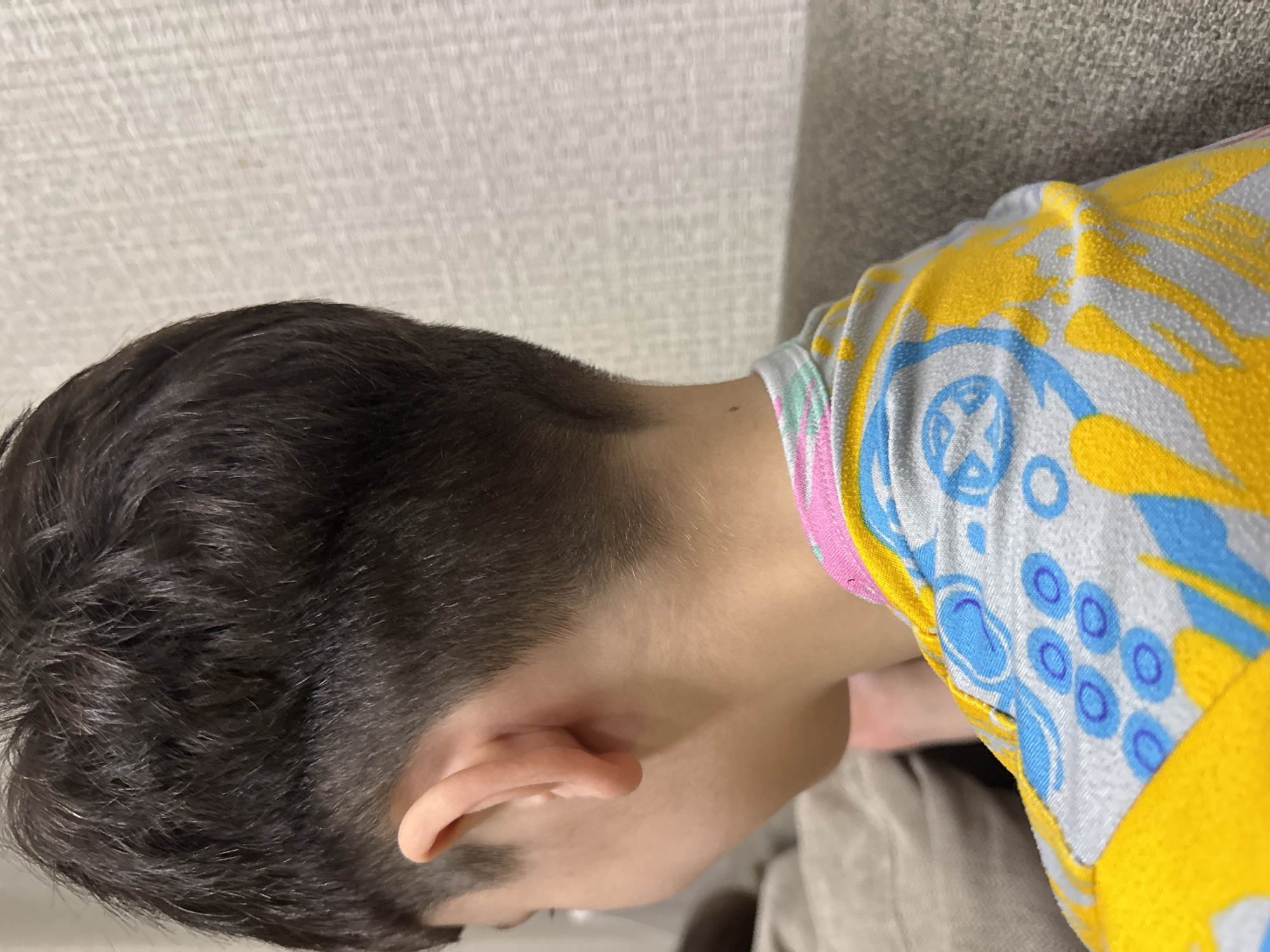 A close-up, side-profile shot of the back of a young person's head and neck is shown. The person has dark, short hair that is styled in a fade on the sides and back. The person is wearing a light-colored t-shirt with a colorful abstract pattern of yellow, blue, and pink. The background is out of focus and appears to be a textured wall and a couch. No specific city or location is identifiable.