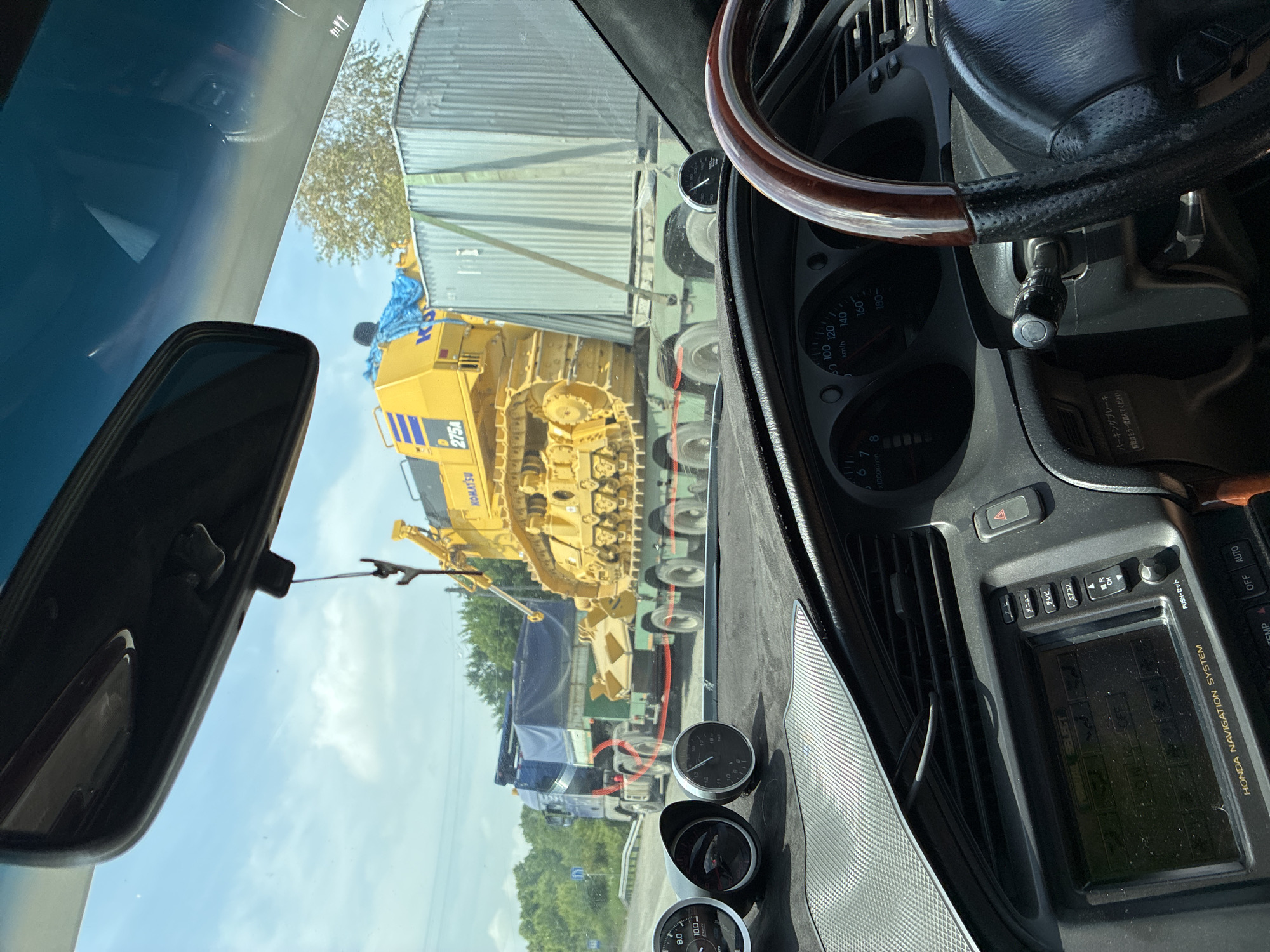 The image shows a view from inside a vehicle, looking forward through the windshield. The vehicle is on a road, and in front of it, a large yellow Komatsu bulldozer is being transported on a flatbed truck. The bulldozer is labeled "275A". Adjacent to the bulldozer on the truck is a large, corrugated metal container. A blue tarp covers part of another truck parked behind the flatbed. The surroundings appear to be rural or semi-rural, with trees visible in the background and a cloudy sky overhead. The interior of the vehicle shows part of the dashboard with gauges and a navigation system. The image was taken in Spassk-Dal'nij, Russia.