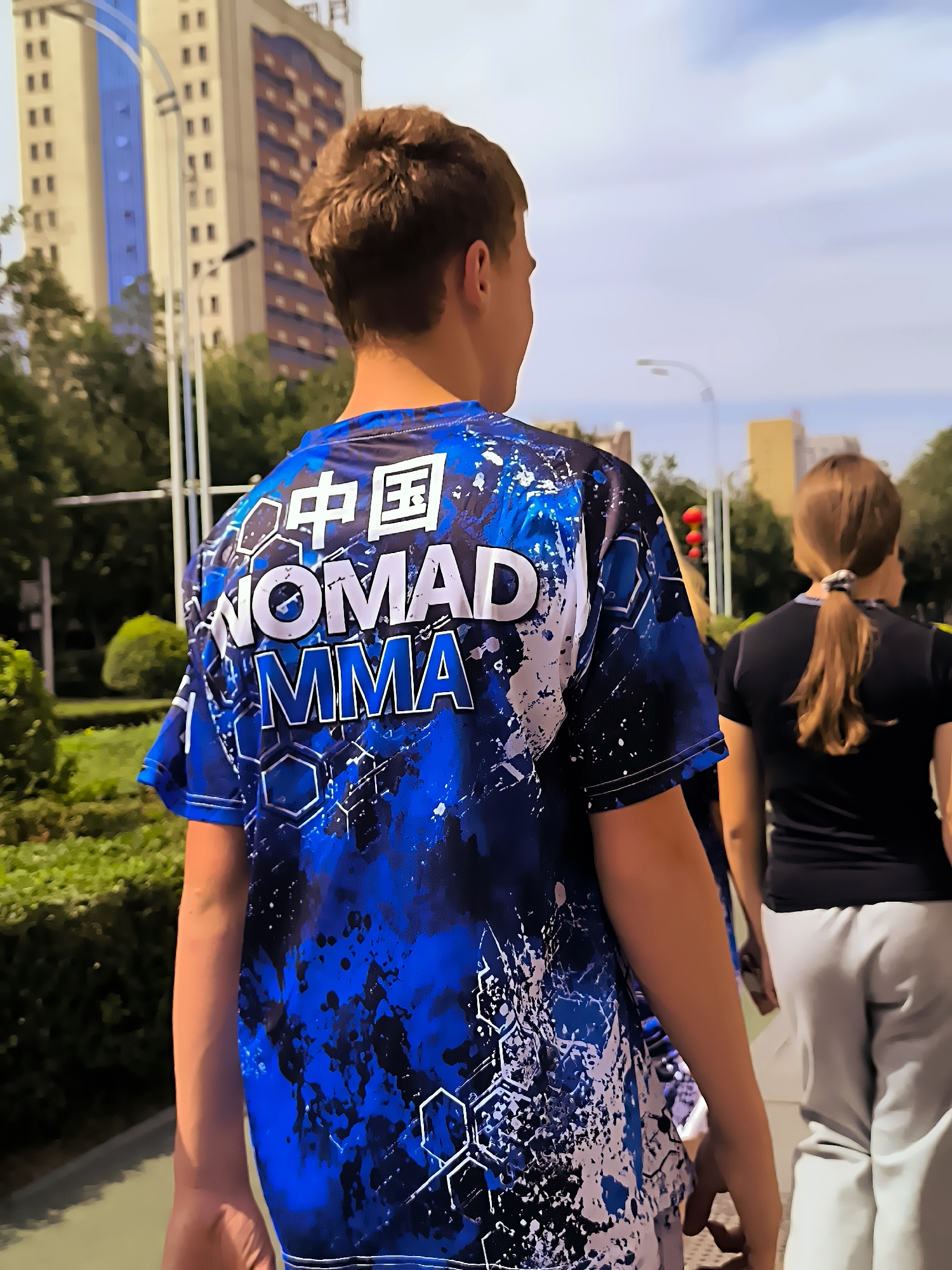 The image shows the back of a young male wearing a blue and white tie-dye t-shirt with black splatters, featuring the text "NOMAD MMA" and Chinese characters. He is walking away from the camera on a sidewalk. Beside him, also walking away, is a young female with brown hair tied in a ponytail, wearing a black t-shirt and grey sweatpants. In the background, there are trees, a lamppost, and tall buildings. One of the buildings appears to be a modern high-rise with a light-colored facade and blue-tinted windows. The setting is outdoors in an urban environment, likely a street or park in Ürümqi, China, indicated by the presence of Chinese characters on the t-shirt and the architectural style of the buildings. The sun is shining, casting shadows, and the sky is visible.
