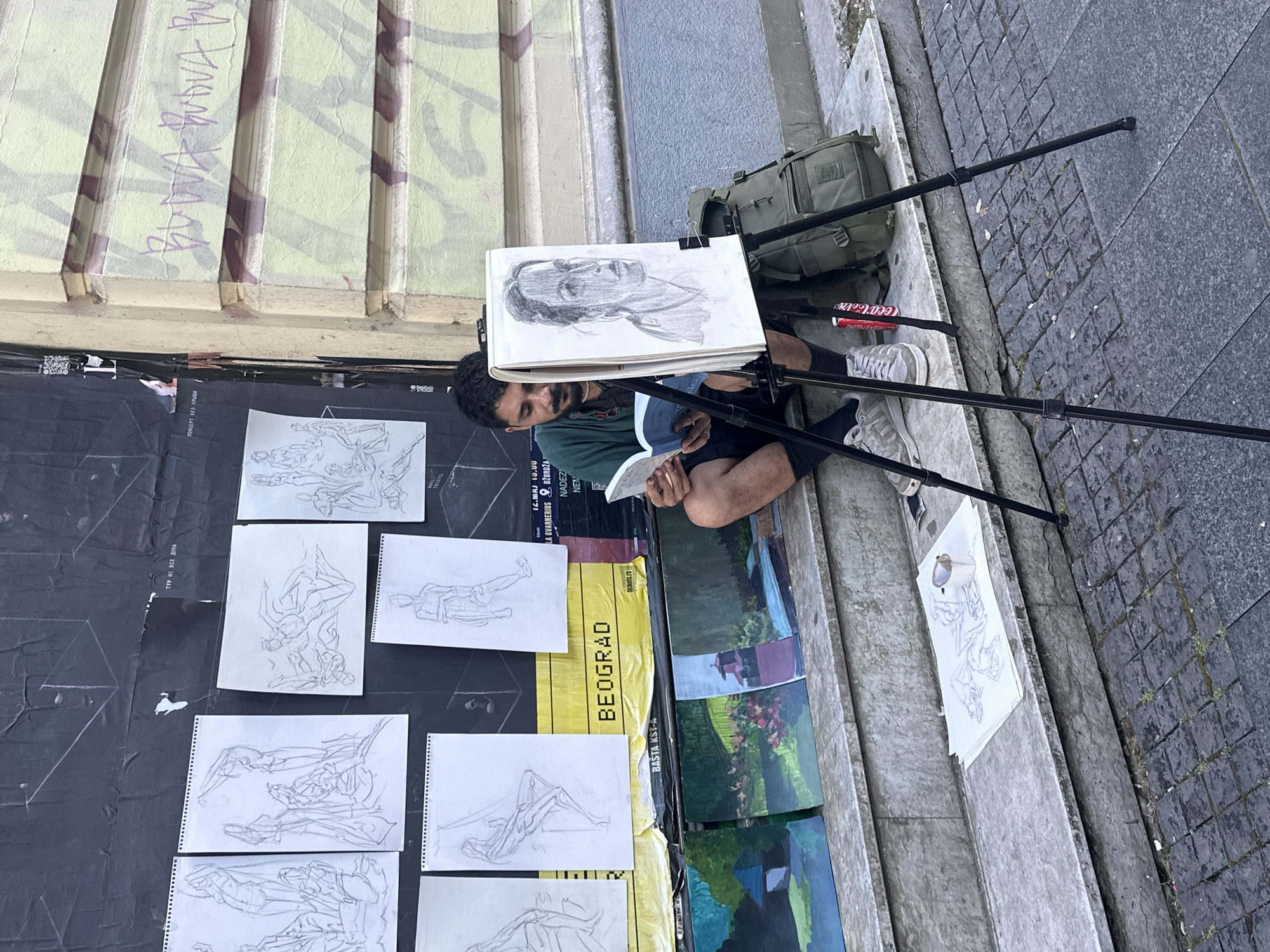 An artist sits on the steps of a building in Belgrade, Serbia, reading a book while his latest portrait is displayed on an easel. The artist is a man with dark hair and a beard, wearing a green shirt and dark shorts. He is sitting on the steps in front of the easel, which holds a charcoal sketch of a woman's face. A backpack and a can of Coca-Cola are next to him. The background shows a wall covered with posters and graffiti. Several other sketches are displayed on the wall and on the steps. The area appears to be a public space, possibly a street or a plaza, with cobblestone pavement in the foreground. The overall scene suggests an urban environment where art is being created and displayed outdoors.