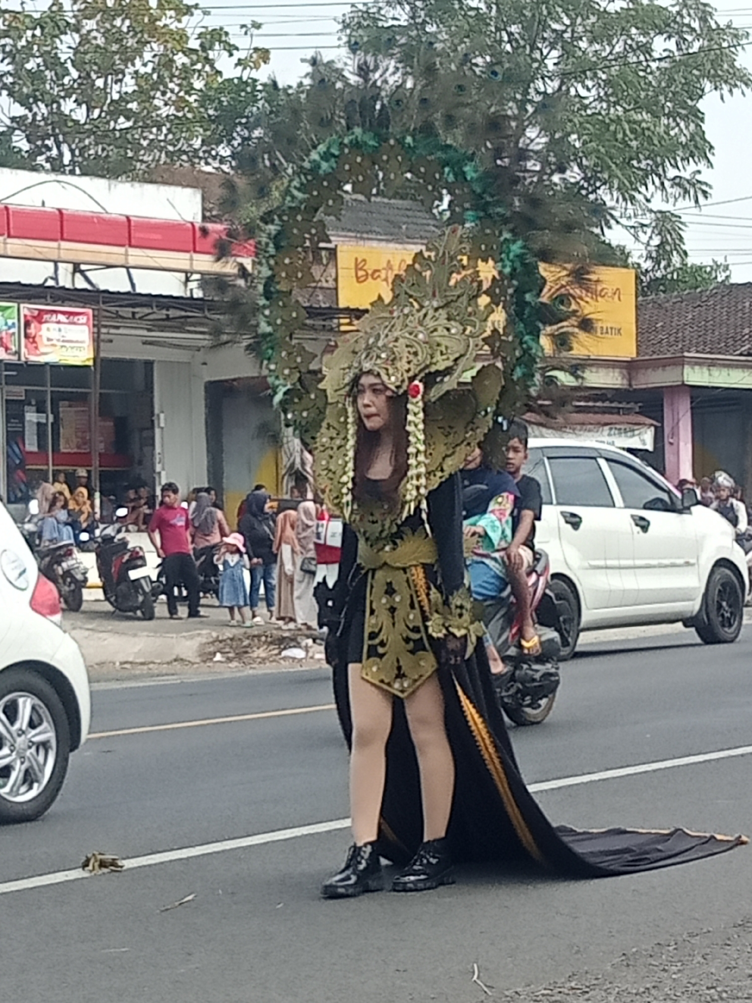 In the city of Urutsewu, Indonesia, a person is captured in a parade, adorned in an elaborate peacock-themed costume. The costume features a large headdress resembling peacock feathers and ornate green embellishments. The individual is wearing a black dress with gold trim, patent leather boots, and is standing on an asphalt road. Behind them, a white car and several motorcycles are visible, with onlookers observing the parade from the roadside. A building with signs can be seen in the background.