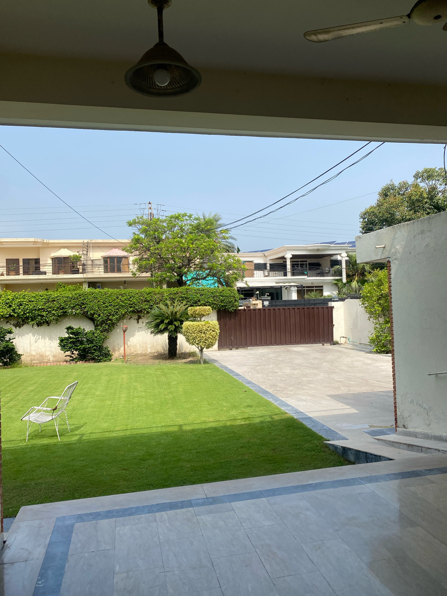 This image depicts the exterior of a residential property, likely in Keer, Pakistan, viewed from an covered patio area. In the foreground, a paved patio with light-colored tiles and a dark blue accent border leads to a well-maintained green lawn. A single white wire chair sits on the lawn. Beyond the lawn, a tall, dense green hedge lines a wall. Behind the hedge, a large brown gate is visible, leading to a concrete driveway. Two residential buildings are visible in the background; one appears to be a multi-story house with a cream-colored facade, topped with solar panels, while another building to the left is also multi-story with similar architectural features. A mature tree with vibrant green foliage stands prominently in the middle ground. The sky is clear and blue, suggesting a sunny day. Overhead, a ceiling fan and a light fixture are visible on the covered patio. Electrical wires are strung across the sky in the background, indicating the presence of utilities.