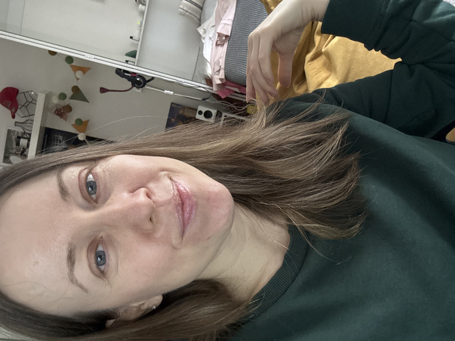 The image captures a close-up portrait of a female subject within an indoor residential environment in Kudrovo, Russia. The subject, a woman with fair skin, blue eyes, and light brown shoulder-length hair, is positioned centrally, gazing directly at the camera with a faint smile and slightly parted, glossy lips. She wears a dark green long-sleeved crewneck top. Her left hand is partially visible in the lower right quadrant, resting on a mustard-yellow fabric surface. The background behind the subject's left shoulder features a white wall with two wall-mounted shelves. The upper shelf displays a red baseball cap, a white vase, and other assorted items. A decorative garland composed of triangular fabric pieces in orange, green, and white is strung across the wall. Below the shelves, a white speaker on a stand and a red and black stick vacuum cleaner are present. Behind the subject's right shoulder, a large mirrored surface reflects a section of the room, revealing a bed with a grey and white striped comforter and pink clothing draped over it, along with a white shelf holding small bottles and dark headphones hanging from a wall hook.
