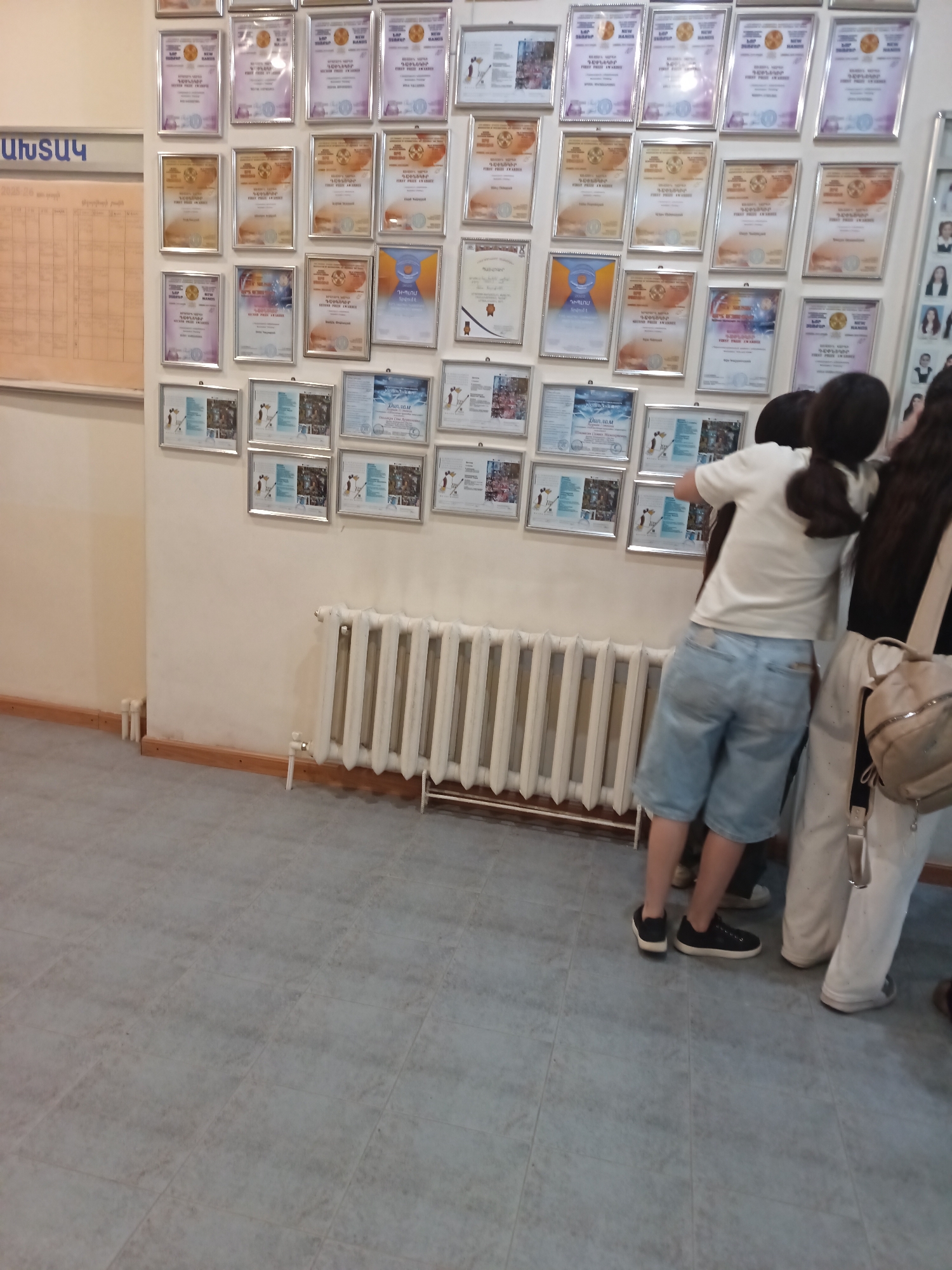 In an indoor setting, likely a school hallway, a wall is adorned with numerous framed certificates. The certificates are arranged in rows and columns, predominantly displaying a gradient of colors from purple to orange, with some featuring circular emblems. A notice board with a grid layout and some text is visible on the left. To the right of the certificates, a white radiator spans across the lower portion of the wall. Three individuals are partially visible in the foreground, their backs to the camera, seemingly observing the certificates. One person is wearing a white t-shirt and blue denim shorts, another a black t-shirt, and a third a black top. The floor is tiled in a neutral gray color.