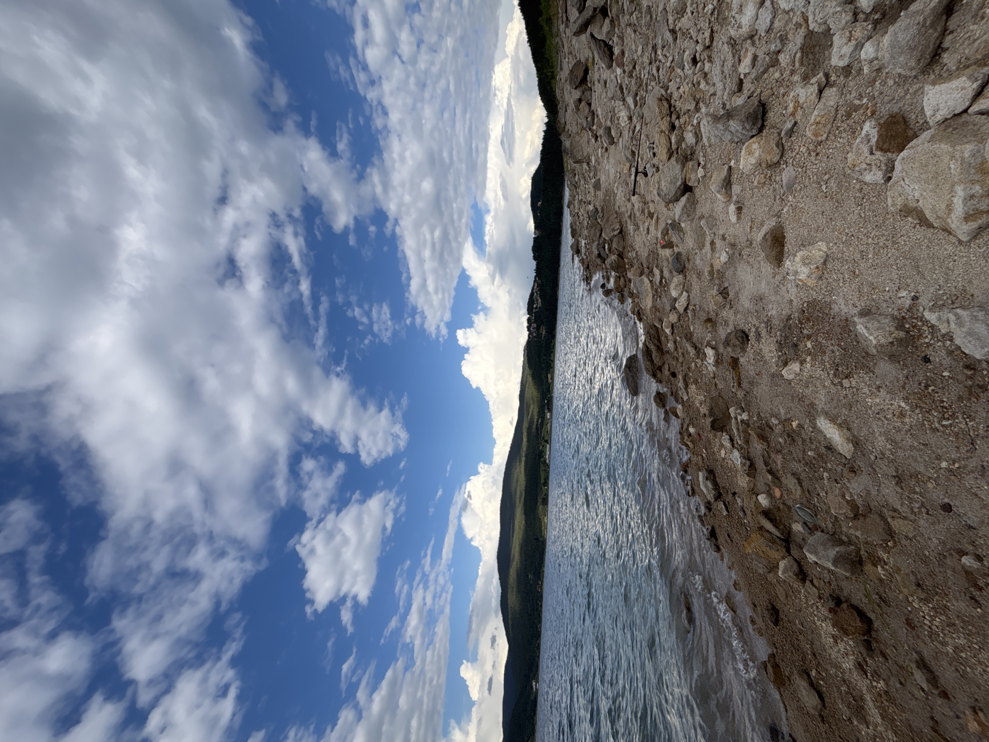 The image shows a rocky shoreline with a body of water extending into the distance. In the background, a tree-covered mountain range is visible under a partly cloudy sky. The foreground is dominated by a rocky beach with various sizes of stones and pebbles. The water appears calm with small ripples near the shore. There are no visible people or animals in the image. The environment suggests an outdoor, natural setting, possibly a lake or reservoir, with a rugged, undeveloped coastline.
