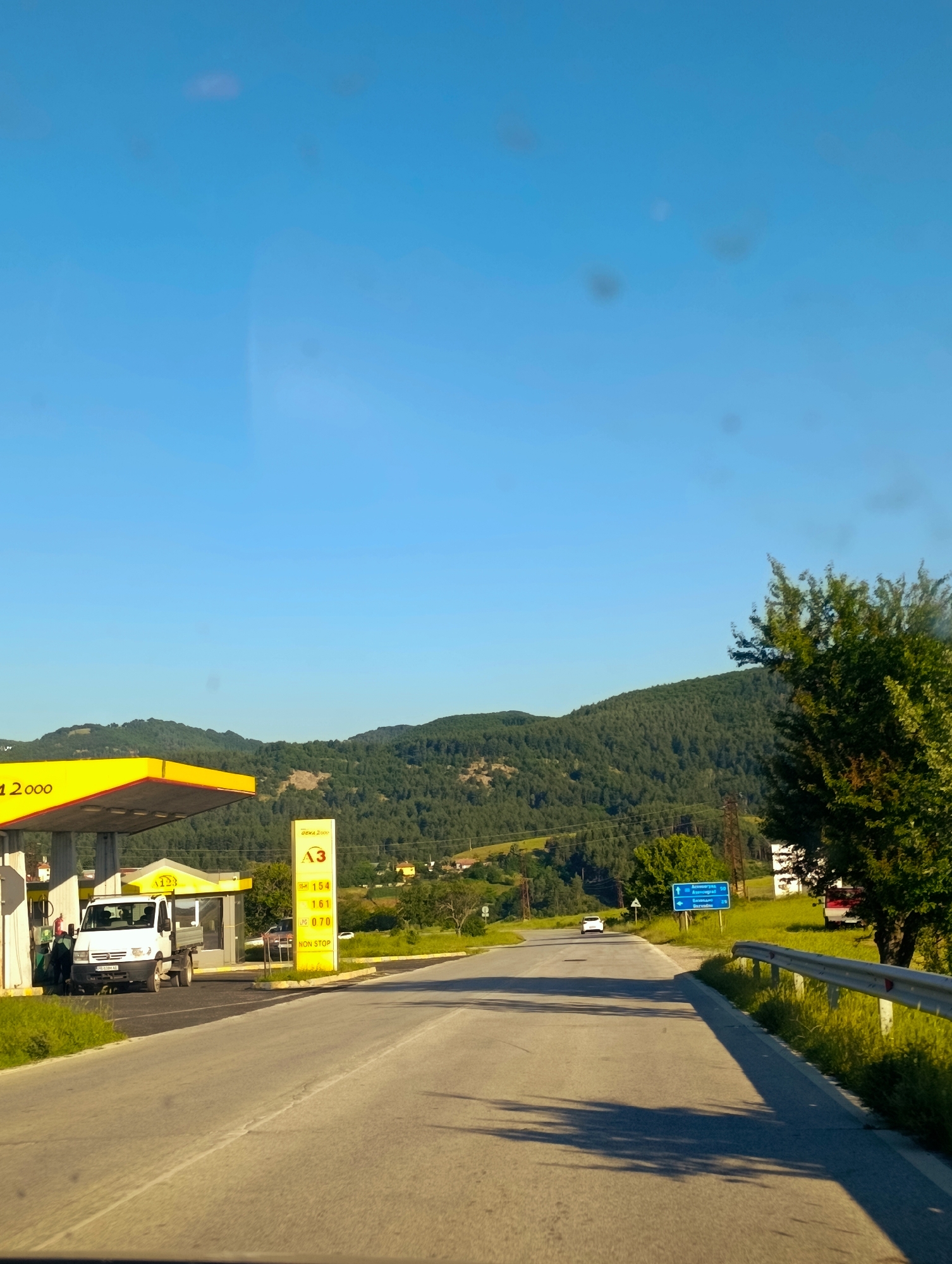 The image captures a scene at a roadside gas station in Komuniga, Bulgaria. The gas station features a yellow canopy over the pumps and a tall vertical sign displaying fuel prices. A white truck is parked at one of the pumps. The road in front of the gas station curves to the right. Beyond the road, a lush green forested hillside forms the background, with scattered buildings visible in the distance. The sky is clear and blue. A blue directional sign with white lettering is posted on the right side of the road.
