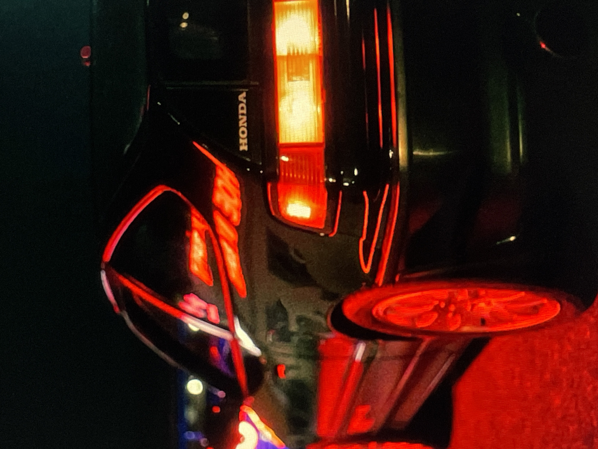 The image depicts the rear of a black Honda vehicle, with the "HONDA" logo visible near the illuminated tail light. The location is a nighttime setting, characterized by dark surroundings and vibrant red lighting, possibly from neon signs or traffic signals reflected on the car's body. The presence of bright lights and reflections indicates an urban environment.