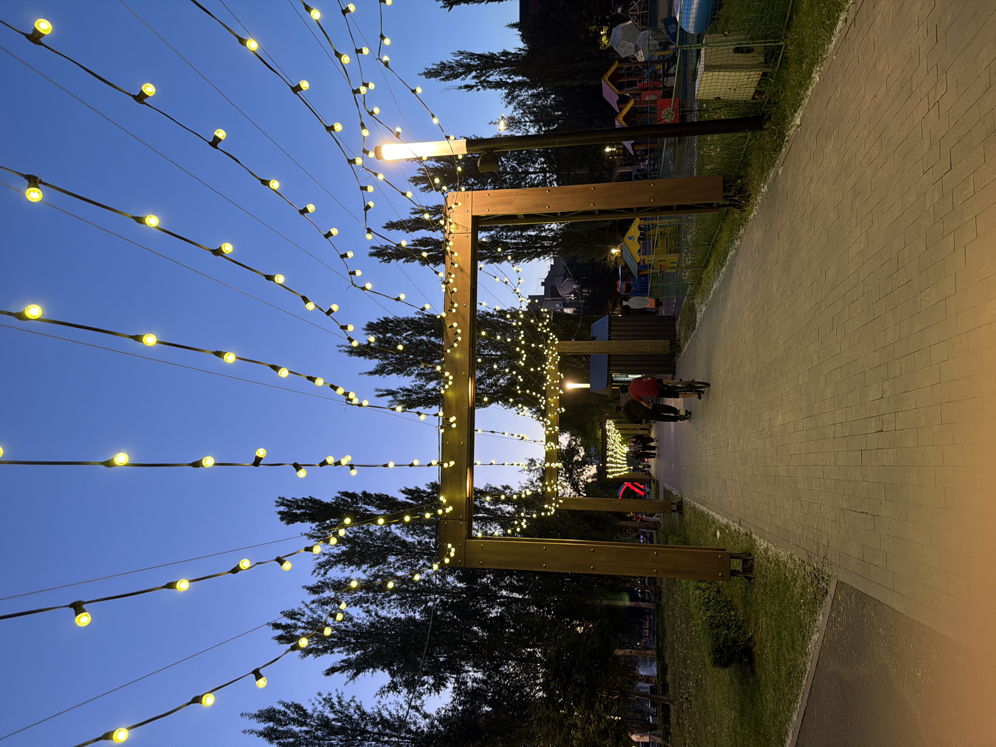 Here is a detailed description of the image:
**Scene:** The image captures a park pathway at dusk, illuminated by strings of lights suspended above. The pathway, paved with rectangular stones, stretches into the distance, lined with grass on both sides. Arched wooden structures support the light strings, creating a tunnel-like effect. In the background, trees are visible, along with what appears to be a playground structure.
**People:** Several people are walking along the pathway. One individual is riding a bicycle in the middle of the shot.
**Objects:** The primary objects in the scene are the strings of lights, the wooden archways, the paved pathway, and the surrounding vegetation. A street light is also present.
**Actions/Interactions:** People are walking and cycling along the pathway, enjoying the ambiance of the park at dusk. The lights illuminate the path, creating a pleasant and inviting environment.
**Environment:** The scene is set in a park, likely within a city. The presence of trees and grass suggests a natural environment within an urban setting. The time of day is dusk, as indicated by the darkening sky and the illumination provided by the lights.
**Location:** The location of the scene is unknown.