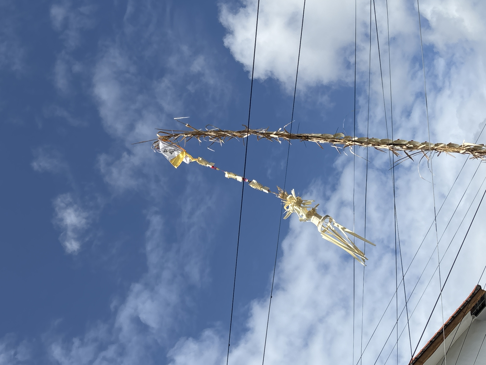 In Kuta Selatan, Indonesia, a traditional Balinese *penjor*, a towering bamboo pole adorned with offerings, is erected against a backdrop of a bright blue sky with scattered white clouds. The *penjor* is decorated with woven palm leaves and what appears to be small offerings wrapped in white and yellow fabric near the top. Power lines are visible traversing the scene horizontally, intersecting the view of the *penjor*. In the lower left corner of the image, the roof of a white building with a terracotta-tiled roof is partially visible. The image is a low-angle shot, emphasizing the height of the *penjor* and the expanse of the sky. No people are visible in the frame.