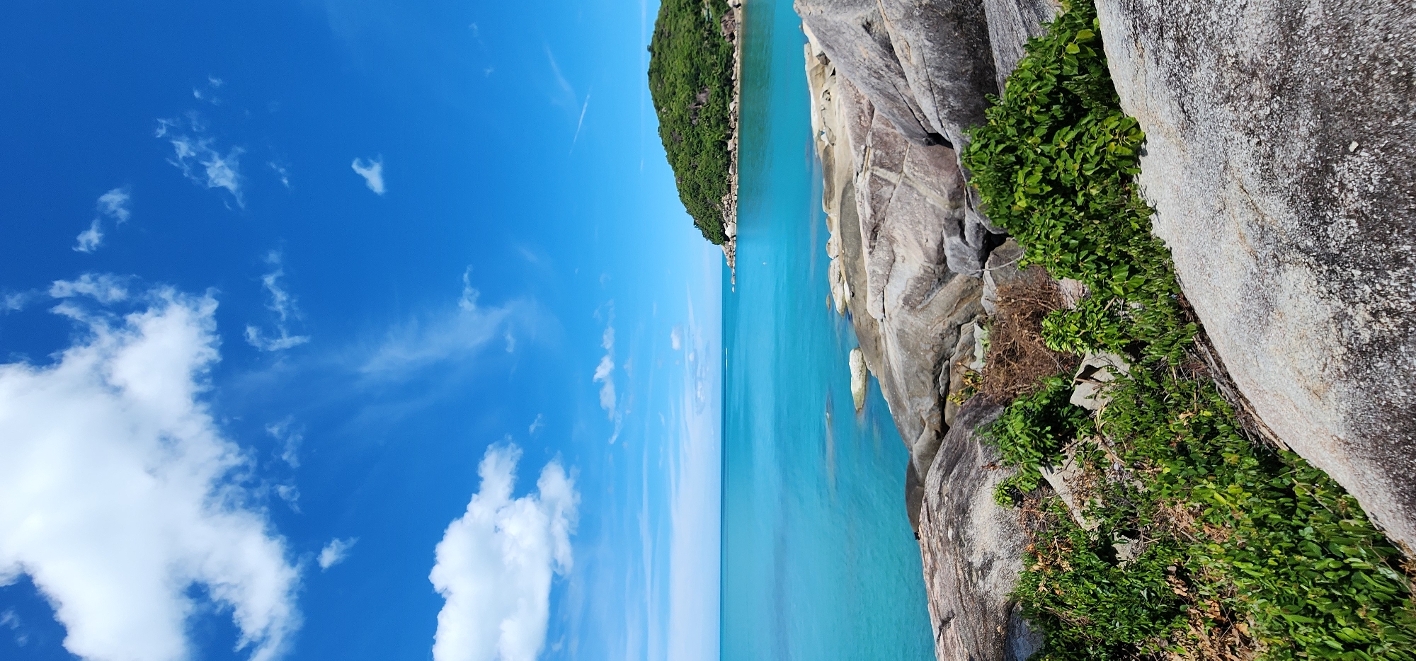 The image depicts a coastal scene in Baan Chaweng Noi, Thailand, under a bright blue sky dotted with white, wispy clouds. The foreground is dominated by large, weathered gray rocks, some of which are partially covered in vibrant green foliage. The rocks transition to smooth, light-colored surfaces with a granular texture in places.
Beyond the rocky shore, the turquoise ocean stretches out, its surface calm and reflective. A low-lying, tree-covered island with a rocky base is visible in the distance, situated to the right of the frame. The vegetation on the island appears dense and lush. The water appears shallow near the shore, with visible submerged rocks and lighter-colored patches indicating sand or coral beneath the surface. The horizon line is clearly defined, separating the sea from the sky. No people or artificial structures are visible in the image. The overall impression is one of a natural, serene tropical landscape.