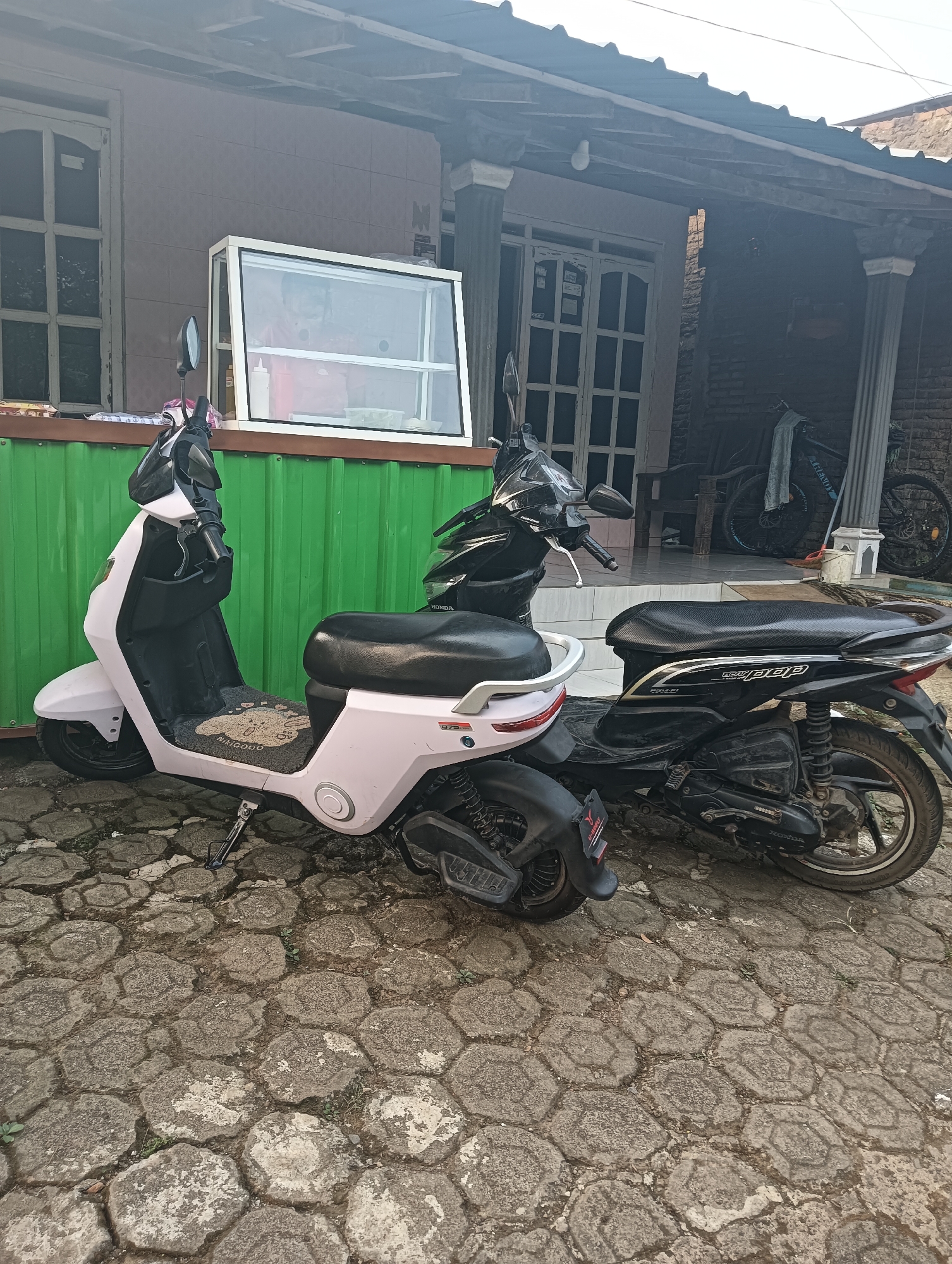 The image depicts an outdoor scene in front of a building, likely a residence or small business, in Cluwak, Indonesia. Two scooters are parked on a textured, hexagonal paved surface. The scooter on the left is a light pink electric scooter, while the scooter on the right is a black Honda scooter. Behind the scooters, a green corrugated metal wall is visible, supporting a glass display case containing various items. The building behind the wall appears to be a house with columns and wooden doors. In the background, a bicycle is leaning against the wall. The overall environment suggests a daytime setting with natural lighting. There are no people visible in the image.