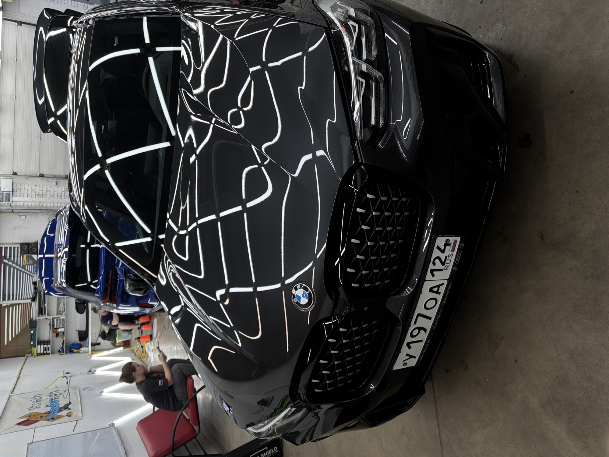 The image shows the front of a black BMW car with a Russian license plate "У 197 ОА 124 RUS", indicating registration in the Krasnoyarsk region of Russia, specifically the city of Krasnoyarsk. The car is parked in what appears to be an indoor garage or detailing shop. The car's surface is highly reflective, mirroring the ceiling lights. In the background, there are at least two other cars, one blue. A person is seen sitting on a red chair in the lower-left corner of the image, possibly using a mobile device. The environment looks like a car detailing or repair shop.