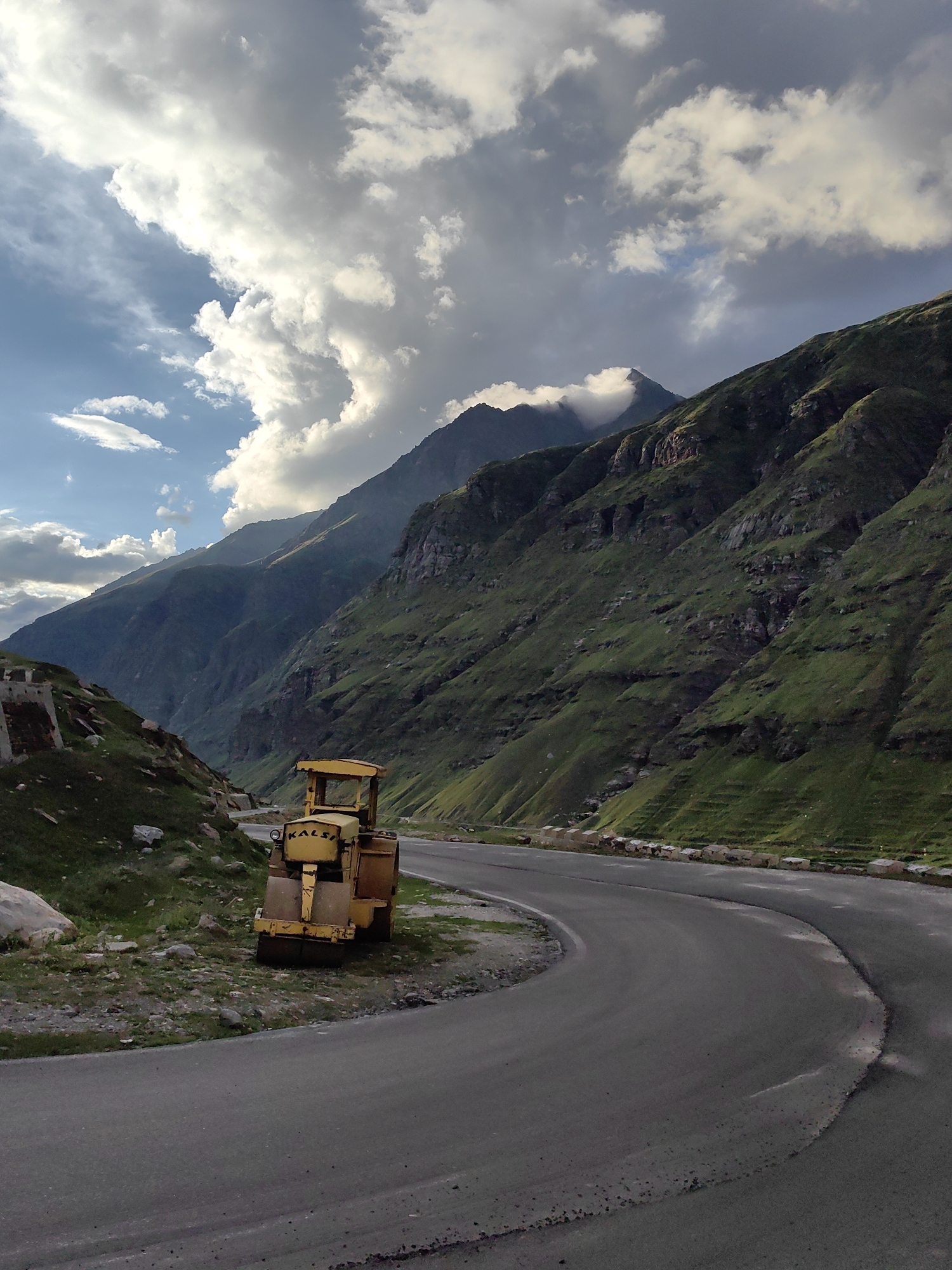 In Yari-Khoksar, India, a yellow road roller sits parked on the grassy shoulder of a winding asphalt road that curves through a dramatic mountain landscape. The road roller, with "KALSI" visible on its side, is positioned in the foreground to the left of the road. Lush green, terraced mountains rise steeply on both sides of the valley, their slopes marked by rocky outcrops and patches of vegetation. The sky above is dynamic, with a mix of bright white, cumulus clouds and darker, more ominous grey clouds, suggesting potential for changing weather. The lighting is soft, casting gentle shadows and highlighting the textures of the mountains and the road. The scene conveys a sense of isolation and rugged natural beauty, with the man-made road and machinery juxtaposed against the grand scale of the Himalayas.