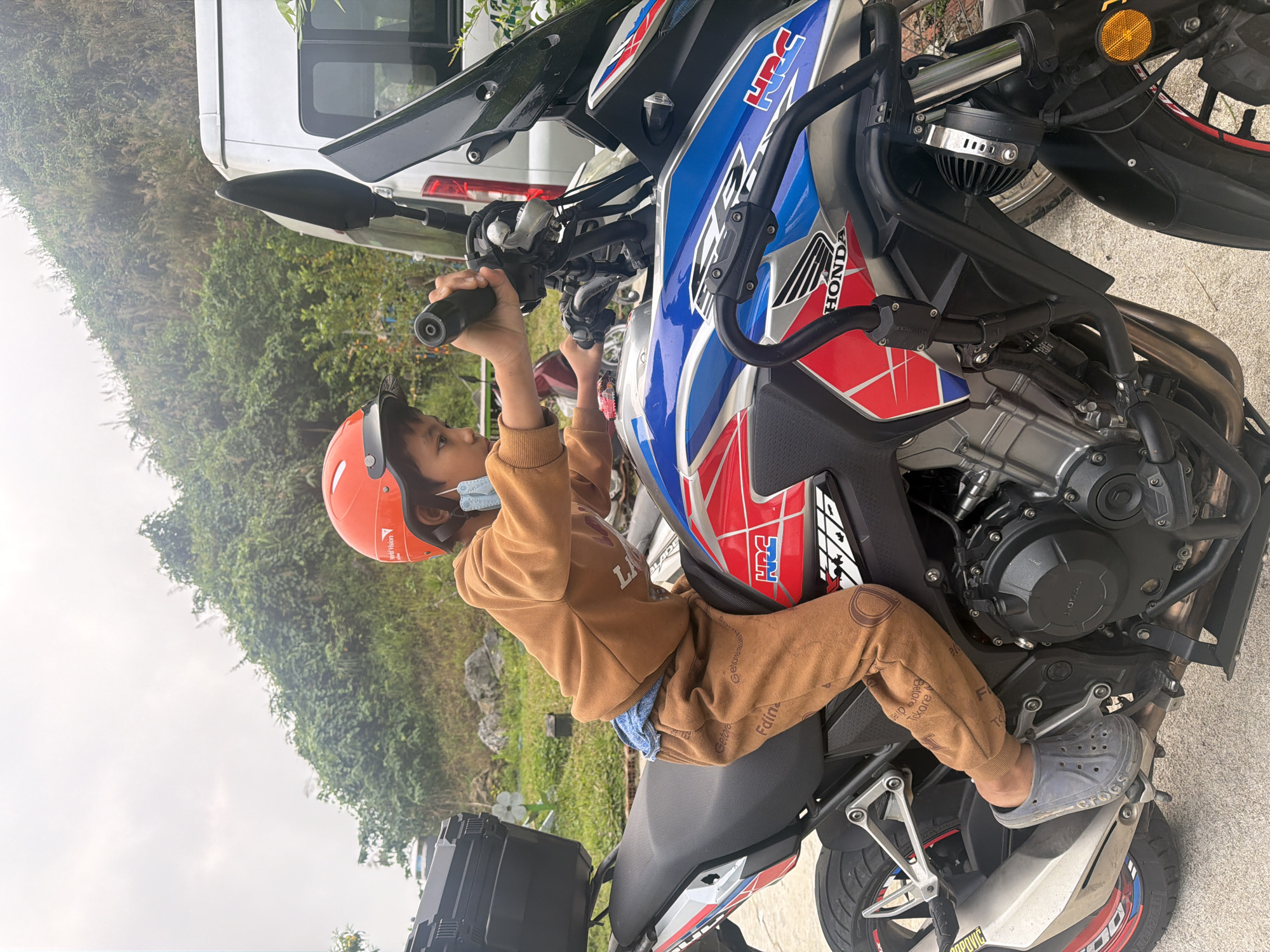 A young child wearing an orange helmet, a brown top, and brown pants is sitting on a motorcycle. The motorcycle is red, white, and blue with the Honda logo. The child is holding the handlebars. The background shows a van, trees, and foliage. The photo was taken outdoors on an overcast day. The location is uncertain and unidentifiable from the image.