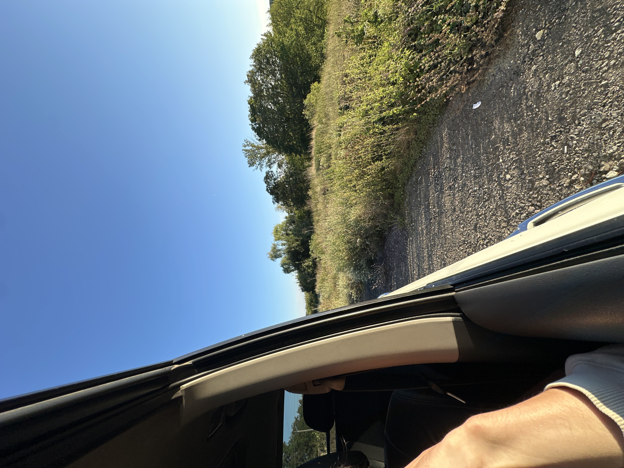 The image shows a view from inside a vehicle, looking out the open door towards a rural landscape under a clear blue sky. The interior of the vehicle is visible on the left, including a black leather seat and a beige pillar. A person's arm and hand are in the foreground on the left, resting on the seat. Outside the vehicle, a gravel road runs alongside a field of tall, green grass and various wild plants. In the background, there are trees and a distant horizon. The scene appears to be a snapshot of a journey through the countryside, possibly near the village of Makiv, Ukraine, given the context. The lighting suggests it is daytime, with the sun casting long shadows.