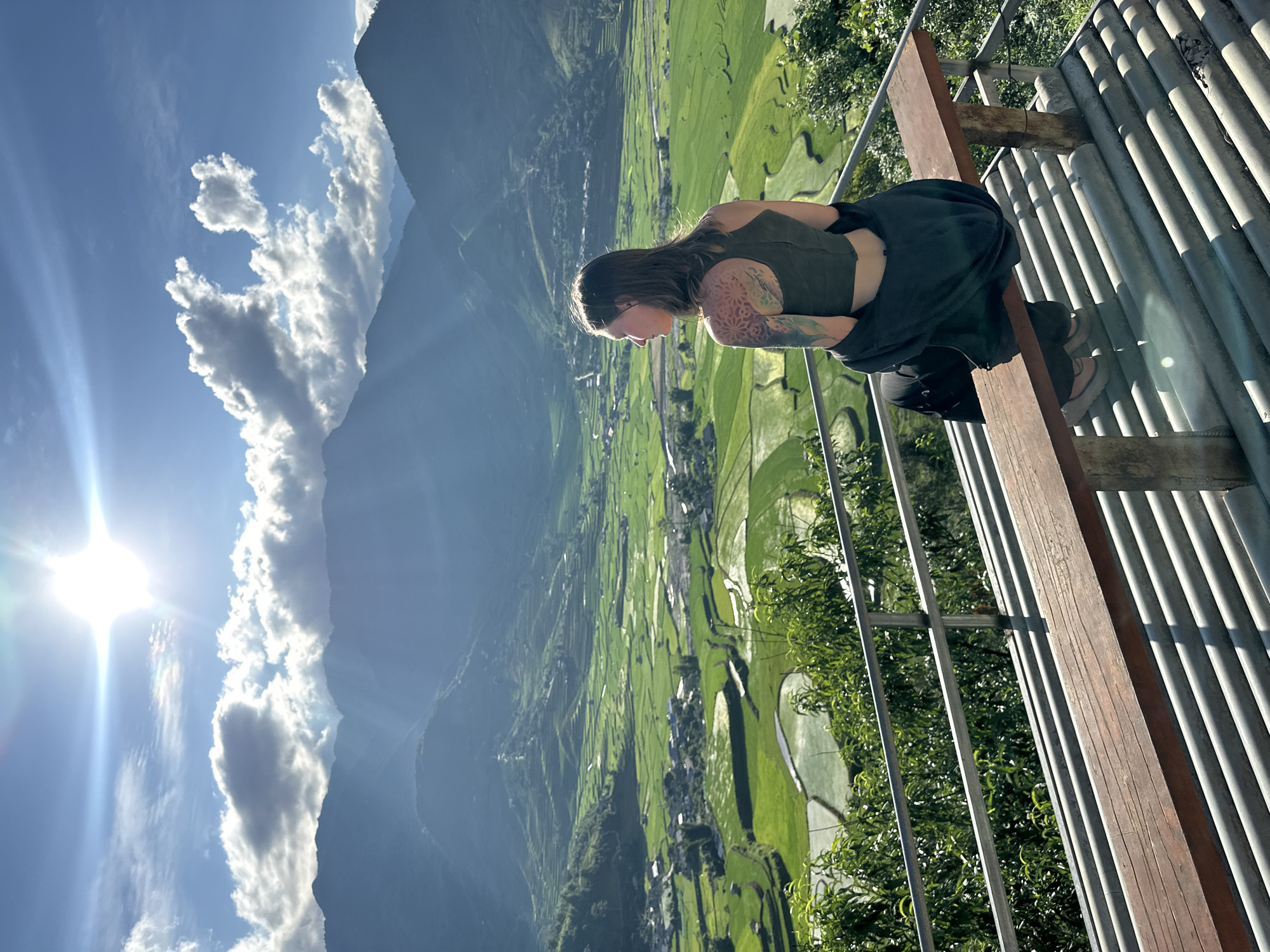 A person is seated on a wooden bench at an elevated viewpoint, overlooking the terraced rice paddies of Tú Lệ, Vietnam. The female subject, positioned with her back mostly to the camera, is looking leftward into the expansive green valley. She wears a dark olive crop top, dark pants, and a dark jacket draped over her lap, revealing a multi-colored, intricate tattoo on her left shoulder and upper arm. The bench rests on a corrugated metal platform, with a two-bar metal railing and green foliage partially obscuring the immediate foreground. Beyond, a vast landscape of vibrant green terraced rice fields with reflective water sections unfolds, interspersed with clusters of village buildings. Distant mountains rise from the valley, layered in a blue haze. The sky is bright blue with scattered white clouds, and a brilliant sun in the upper right casts a strong lens flare and sunbeams across the scene.