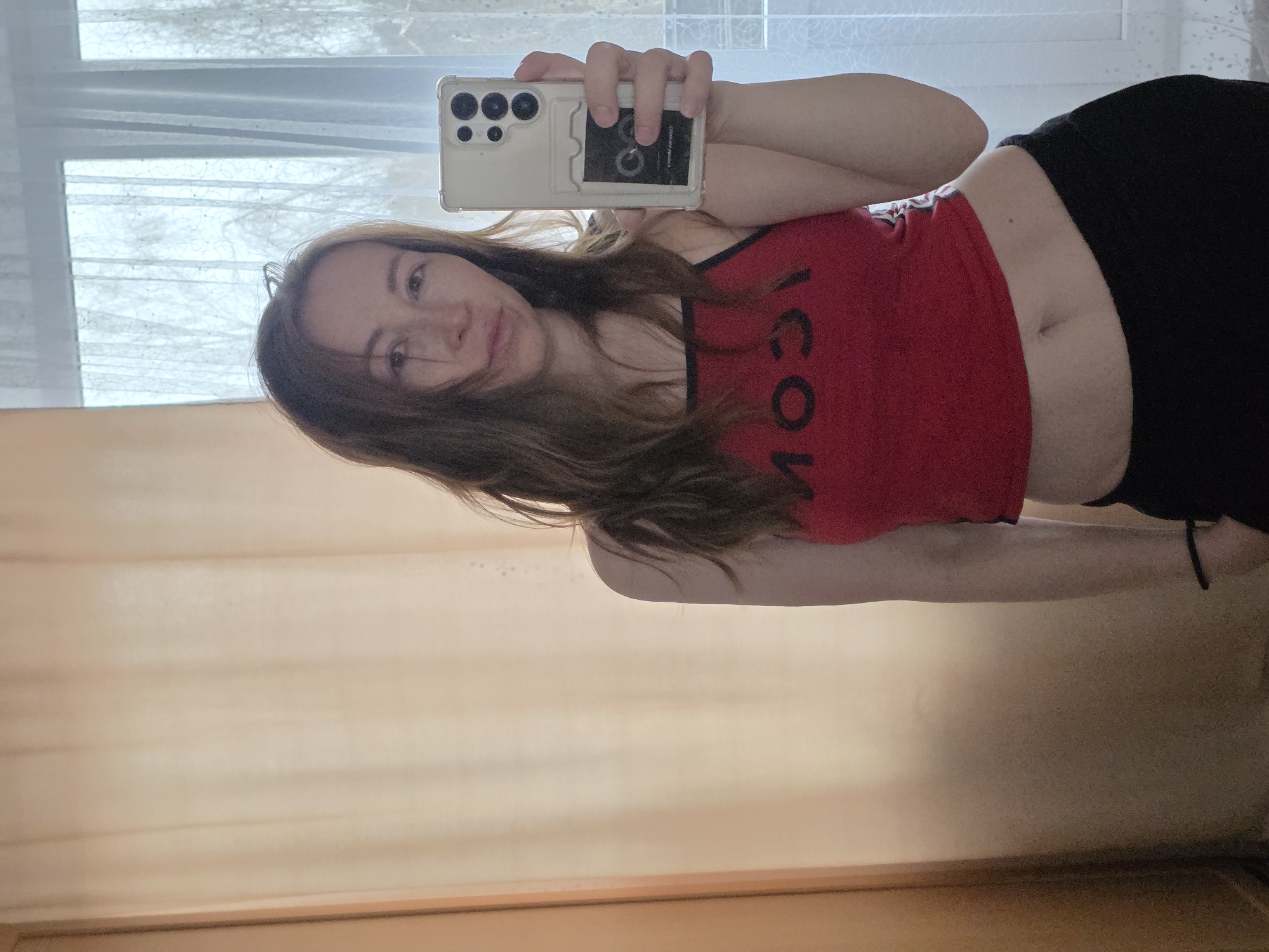A woman is taking a selfie in what appears to be an indoor setting. She is holding a smartphone with multiple camera lenses in her right hand, which is positioned above her head and slightly to the right. The smartphone has a transparent case with a black card partially visible behind it. The woman is wearing a red crop top with black trim and the word "ICON" printed in black on the front. She is also wearing black pants. Her brown hair is partially covering her face.
Behind her, there is a window covered by a sheer curtain, and to the left, there is a wall with what looks like a light-colored curtain or panel. The lower portion of the image reveals a wooden or laminated surface, suggesting the presence of furniture or flooring.
Without further information, the specific location within Chaikovsky, Russia, cannot be determined.