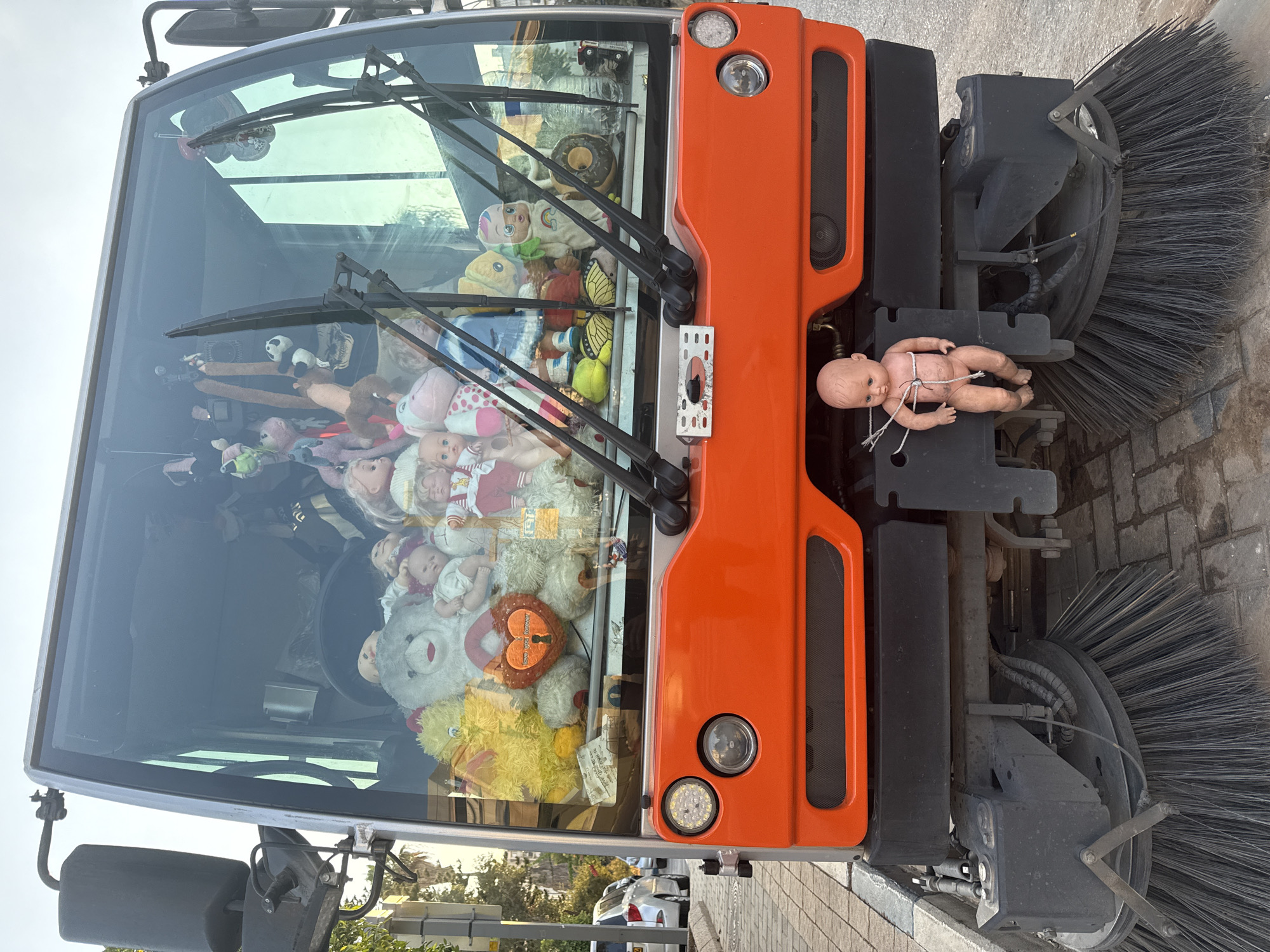A street sweeper vehicle, a Hako Citymaster 1600, is parked on a sidewalk in Tel Aviv, Israel. The vehicle's cabin is filled with stuffed animals and dolls, creating an unusual and eclectic display. A doll is hanging from the front of the vehicle, tied by string. The background shows a street scene with trees and other vehicles.
