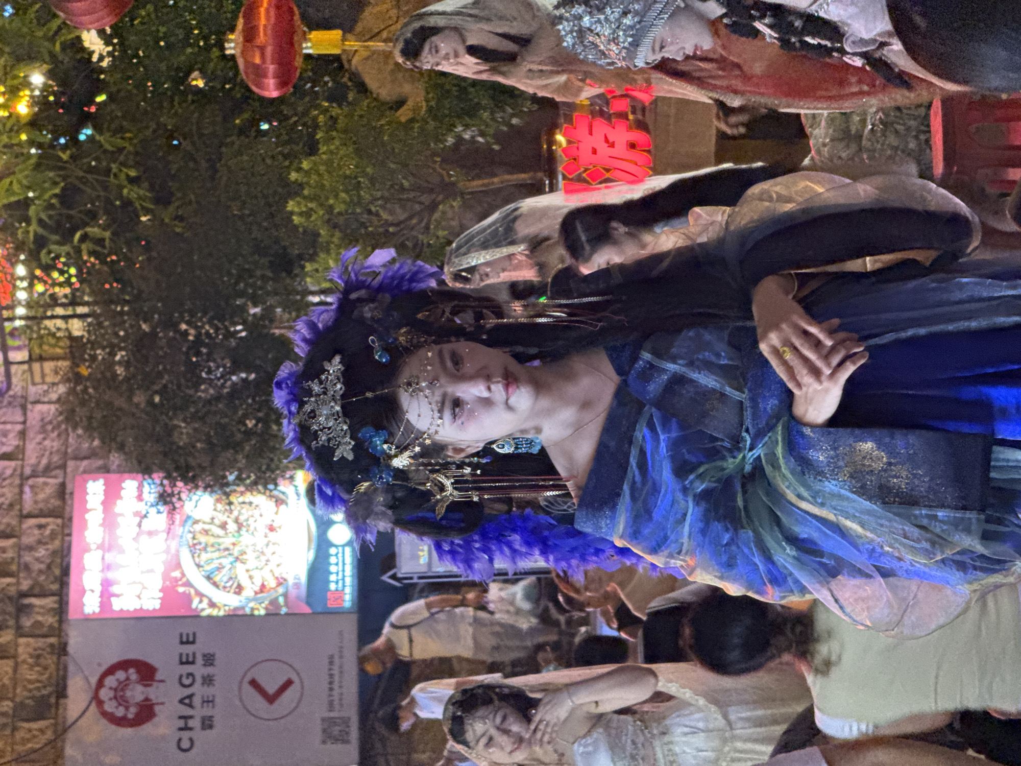 In Yuzhong District, China, a group of women dressed in elaborate traditional costumes are gathered outdoors at night. The central figure, a young woman, wears a striking blue and gold robe with intricate embellishments and a large, ornate headdress adorned with purple feathers and silver accents. Her face is painted with glittery makeup. To her left, another woman in a white, lace-trimmed gown poses with her hand near her face. In the background, several other women in similar attire, some wearing veils or head coverings, are visible. The scene is lit by ambient light and decorative red lanterns. A brick wall forms the backdrop, and a sign with Chinese characters and a logo is partially visible to the left. The overall atmosphere suggests a cultural performance or festival.