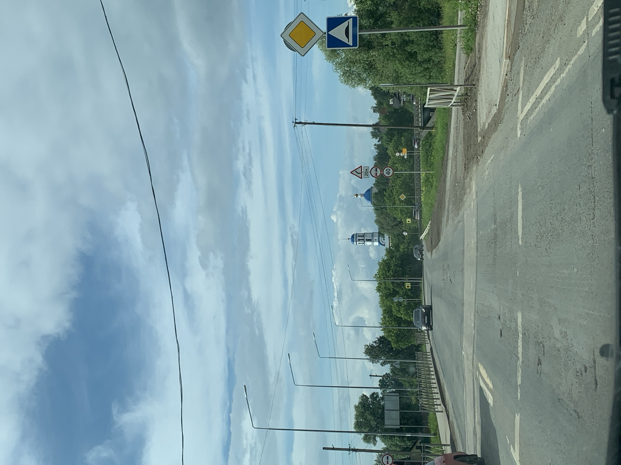 The image captures a view from a vehicle's perspective, looking down a paved road in Orda, Russia. The road is lined with streetlights on the left and features visible road markings, including solid white lines and dashed white lines indicating lanes. The road surface appears worn and has several potholes.
On the left side of the road, a metal railing separates the road from a sidewalk. Beyond the railing, there are trees and a building with a red roof. Further down the road, a cluster of traffic signs is visible. These include a yellow diamond-shaped sign, a blue square sign with a white triangle indicating a speed bump, and a circular sign with the number 40 inside a red circle, suggesting a speed limit.
In the background, a prominent white church with blue domes stands against a cloudy sky. Trees and vegetation are abundant on either side of the road, creating a rural or suburban setting. Several vehicles are present, including a red car partially visible on the left, a dark-colored car in the middle of the road, and other cars in the distance. The sky is partly cloudy with a mix of blue sky and white clouds. The overall scene depicts a typical day on a road in a Russian town.