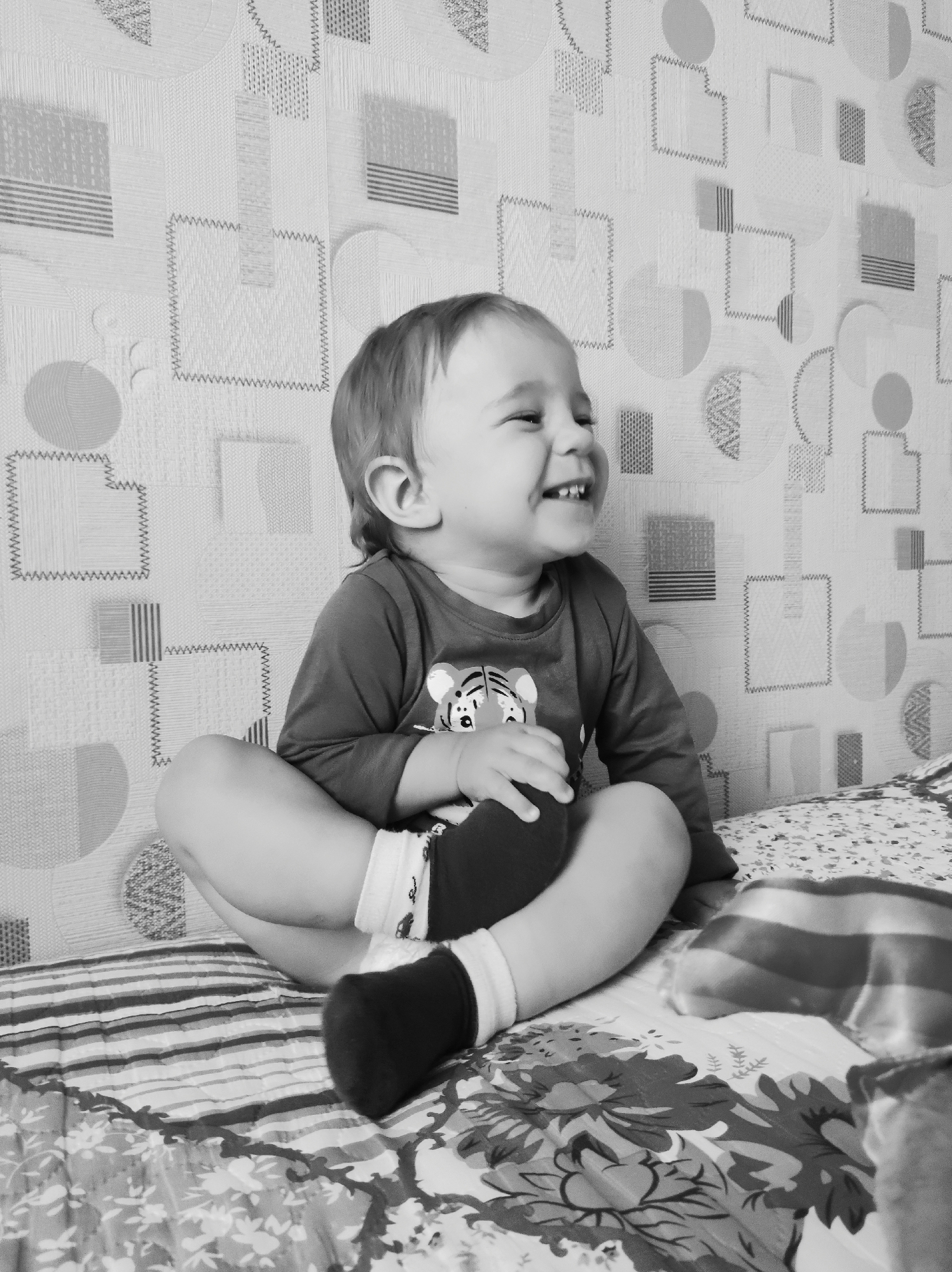 A black-and-white photograph depicts a young child, likely a toddler, sitting cross-legged on a bed. The child is wearing a long-sleeved shirt with a tiger graphic and one black sock. The child is smiling broadly, looking to the side, with their mouth open showing their teeth.
The bed is covered with a floral patterned sheet, and a striped object is partially visible in the lower right corner. The wall behind the child has a patterned wallpaper with geometric shapes.
Based on the information that this image was taken in Chaikovsky, Russia, the photograph likely depicts a scene in a home in that city. There is no other information provided to give additional context.