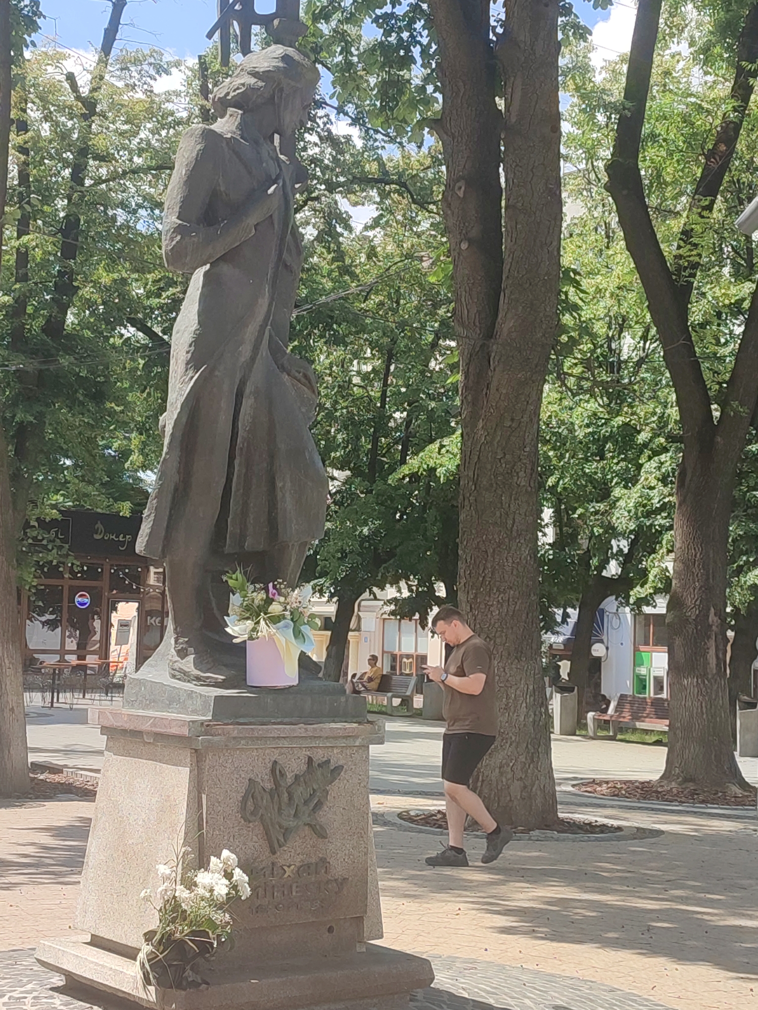 The image shows a bronze statue of a woman in a long dress, standing in a public square. The statue is on a stone pedestal, with a bouquet of flowers at its base. A man in a brown t-shirt and black shorts is walking past the statue, looking at his phone. The background shows trees, buildings, and a few people sitting on benches. The statue is located in the city of Chernivtsi, Ukraine, specifically in the central square, which is a pedestrian zone. The statue is of Olga Kobylyanska, a prominent Ukrainian writer.