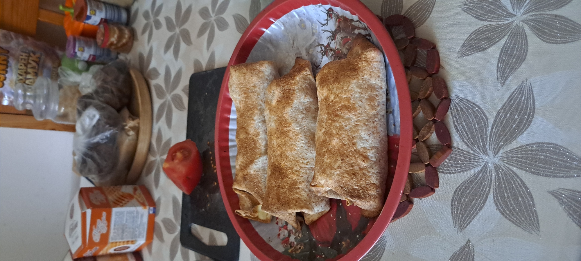 The image shows a close-up, eye-level shot of three golden-brown wraps or burritos arranged on a red tray lined with a festive napkin featuring deer. The wraps are presented on a table with a light-colored tablecloth patterned with stylized grey flowers. To the left of the tray, a wedge of red tomato sits on a black cutting board. In the background, various food items and containers are visible, including a cardboard box with orange and white packaging, a glass jar filled with what appears to be cereal or grain, and other jars and containers with colorful labels. The overall setting suggests a domestic kitchen or dining area. The image does not contain information that allows for specific location within the city of Burgas, Bulgaria.