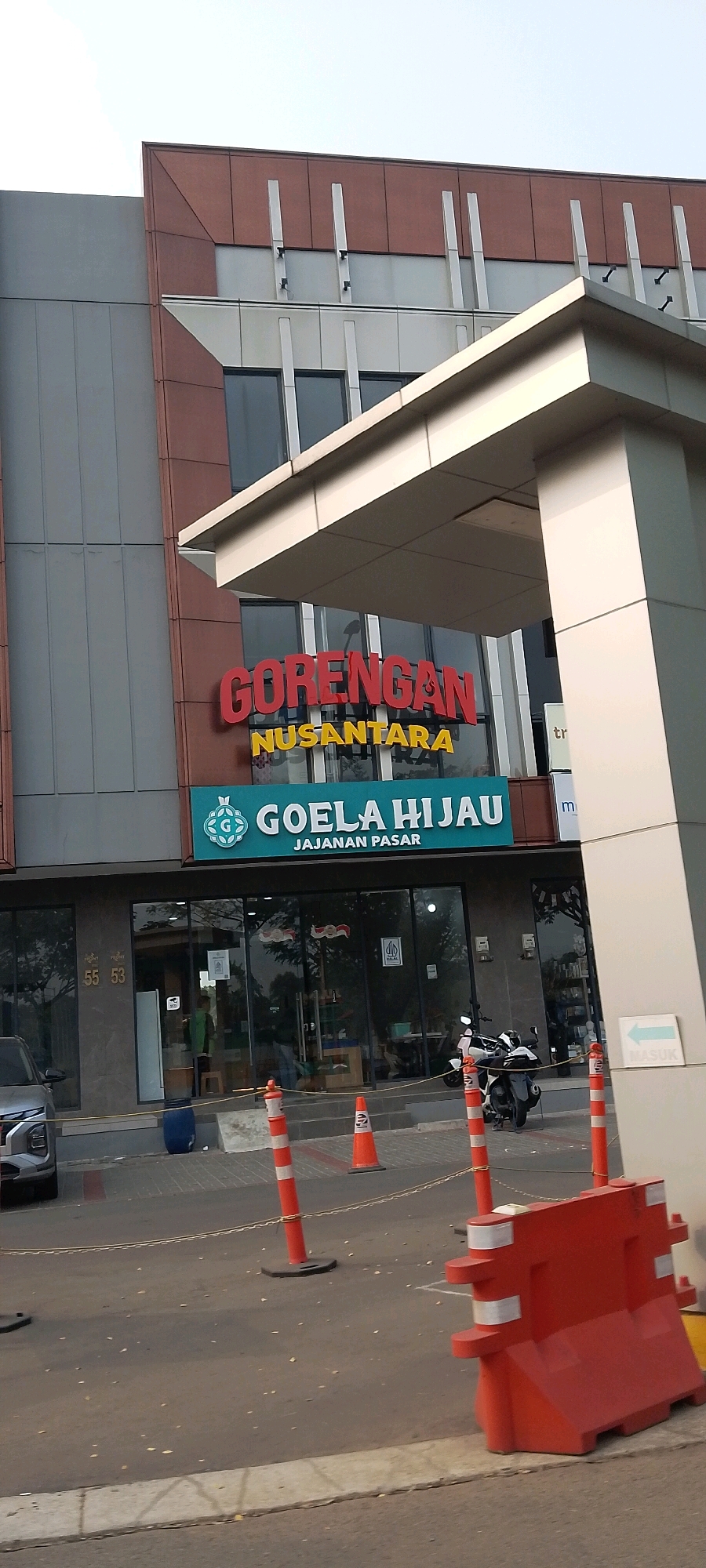 The image shows the exterior of a commercial building in Gunung Putri, Indonesia. The building has a modern facade with dark grey and reddish-brown panels. Two prominent signs are visible above the entrance: "GORENGAN NUSANTARA" in large red lettering and "GOELA HIJAU JAJANAN PASAR" in white lettering on a teal background, with a circular green logo. The entrance features large glass doors. In front of the building, orange traffic cones and a red plastic barrier are present on the asphalt. A silver SUV is parked on the left, and a motorcycle is parked near the entrance. A blue barrel is located to the left of the entrance. A sign with an arrow pointing left and the word "MASUK" (Indonesian for "enter") is attached to a pillar on the right side of the building's overhang. The sky is bright, suggesting daylight.