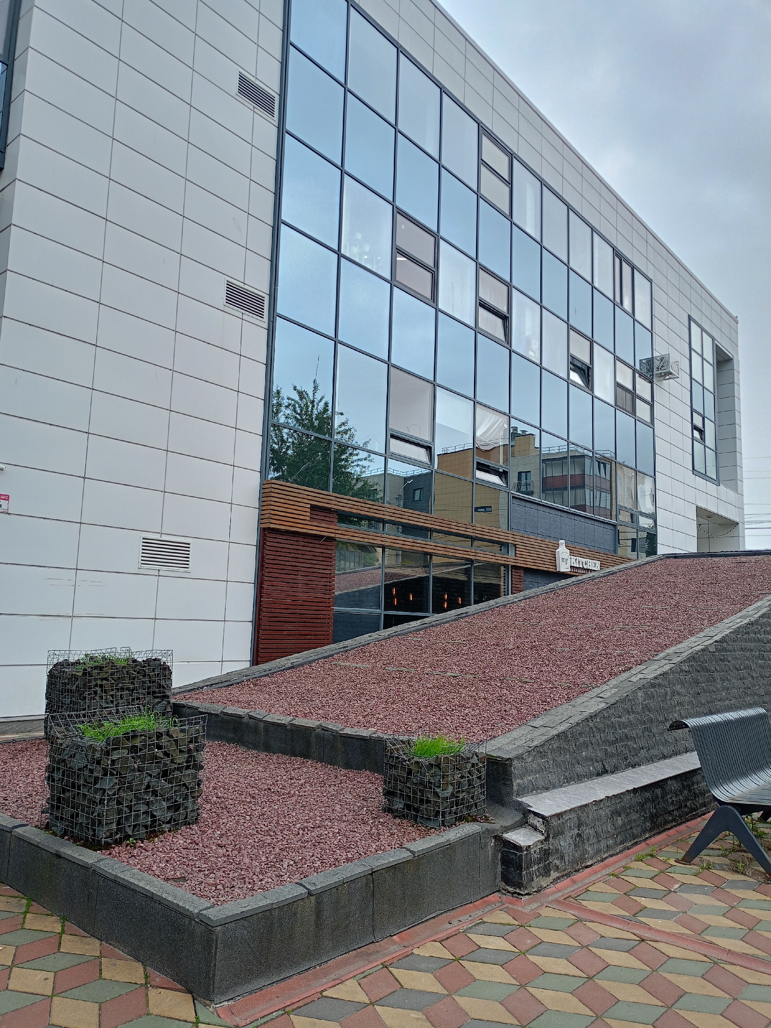 This image depicts the exterior of a modern building with a large, reflective glass facade. The building is multi-story and appears to be made of white tiled panels on the left side and the glass on the right. In the foreground, there are landscaped areas with gravel and gabion planters filled with stones and some greenery. A dark metal bench is visible on the right. The ground is paved with interlocking geometric tiles in shades of red, yellow, and green.
The scene is set outdoors, and the sky appears overcast, suggesting a cloudy or possibly rainy day. The reflections in the glass windows show other buildings and trees, providing some context of the surrounding environment. There is no clear activity or event taking place; the image captures a static view of the building's exterior and its immediate surroundings.
Notable details include the contrasting materials of the building's facade (tile and glass), the angular design of the landscaping, and the patterned paving stones. A small white sign with text is visible on the building above the lower entrance, but the text is too small to be legible in the provided image. Another sign, appearing to be a red warning sign, is mounted on the white tiled wall on the far left. There are also ventilation grilles visible on the white tiled section of the building. The overall impression is one of contemporary architecture and urban landscaping.