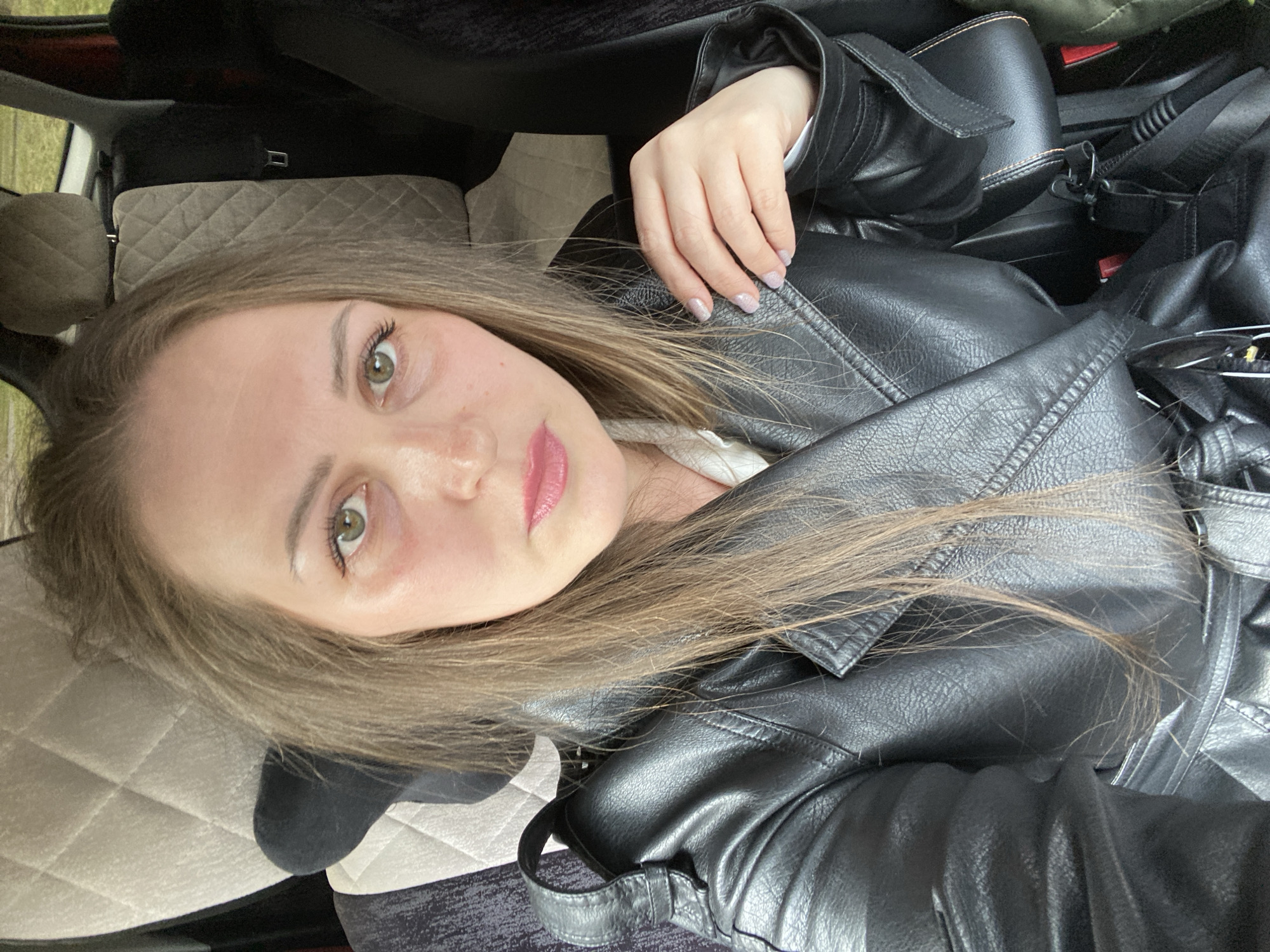 A woman with light brown hair and green eyes is lying down, facing the camera. She is wearing a black leather jacket. She has lipstick on. The background shows the interior of a car, including a headrest and the seat, suggesting she is in a vehicle. The location is Марковский, Russia.