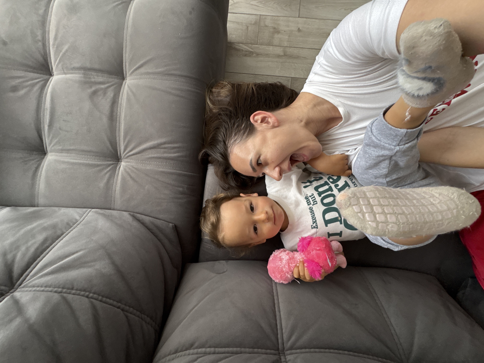 A woman and a child are lying on a gray, cushioned sofa. The woman has dark hair and is wearing a white t-shirt. Her mouth is open, and she appears to be smiling or laughing. The child is wearing a white t-shirt with text and gray pants. They are holding a pink stuffed animal. The background suggests an indoor setting with light-colored wooden floors. There is no information that suggests that the photo was taken in Sochi, Russia.