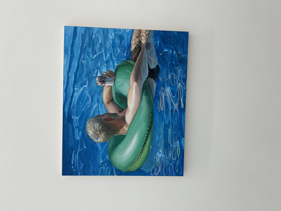 A framed painting hangs on a white wall. The painting depicts a man in a green inner tube floating in blue water. He has short, light-colored hair, and is holding a can of Diet Coke. The water around him is painted with swirling patterns of blue and white, suggesting movement and light reflecting on the surface. The man's upper body is visible, and he appears relaxed and comfortable in the water.
